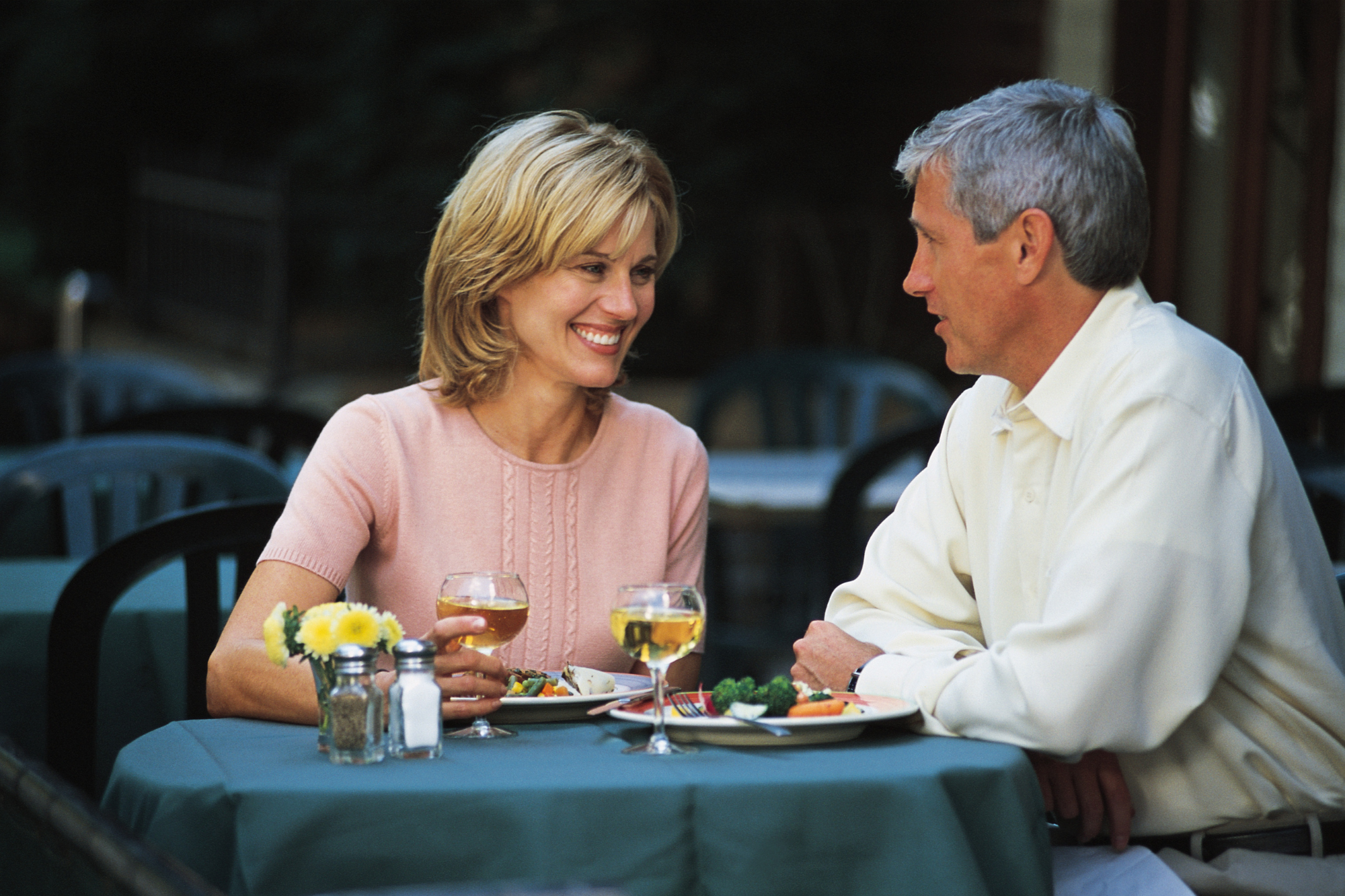 A man and a woman sitting at an outdoor table, smiling and enjoying a meal together, with wine glasses and a plate of food.