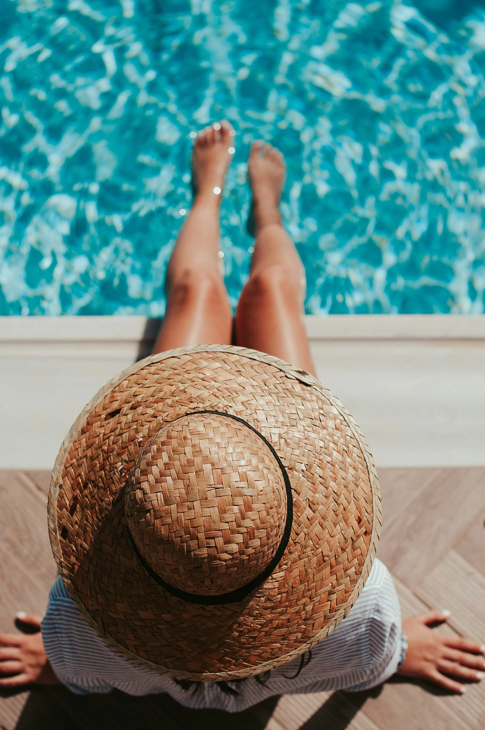 A person sitting by a pool wearing a straw hat, with legs submerged in the water, as seen from above.