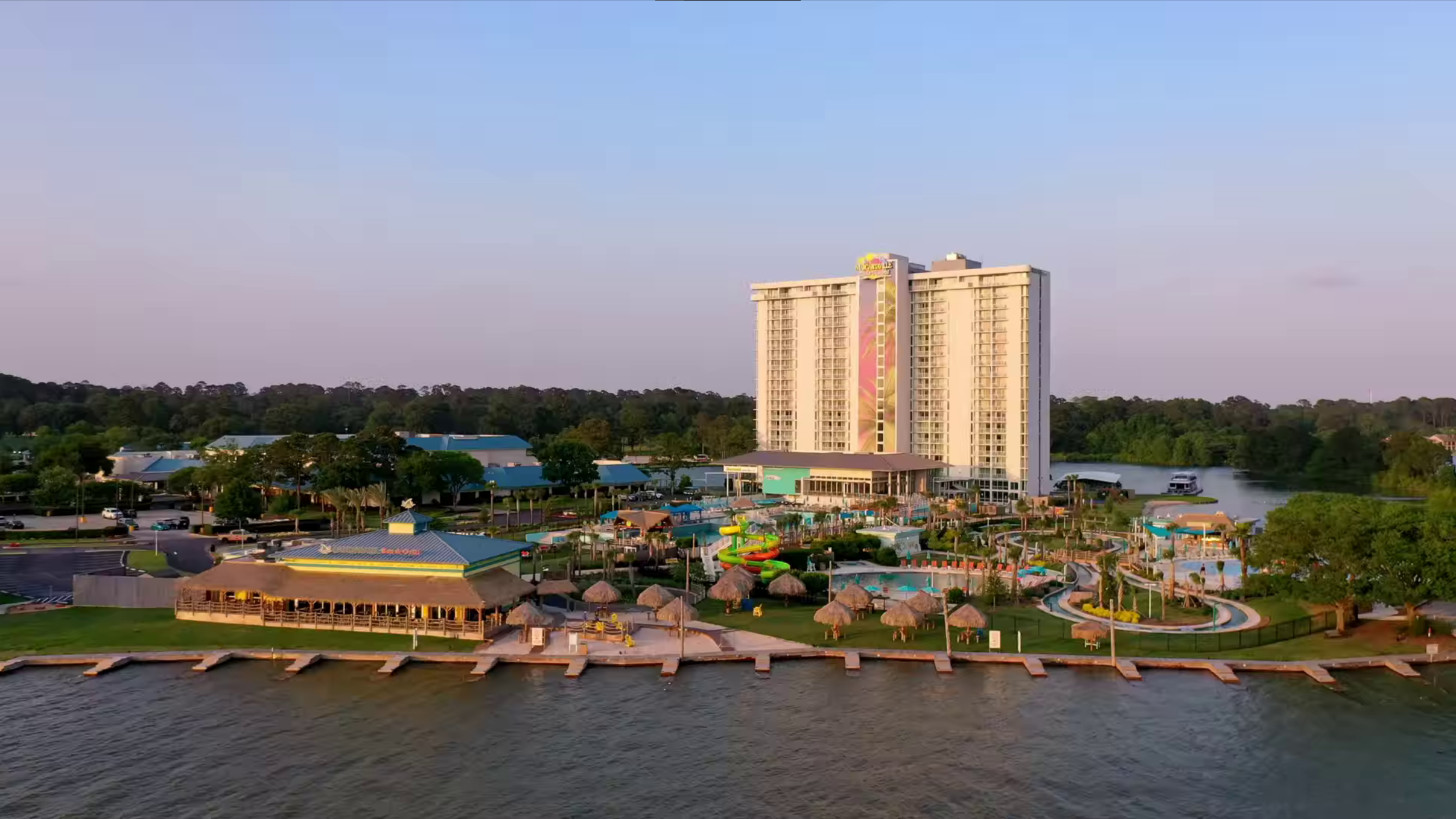 High-rise hotel with colorful murals, water slides, and a lazy river at a lakeside resort during sunset.