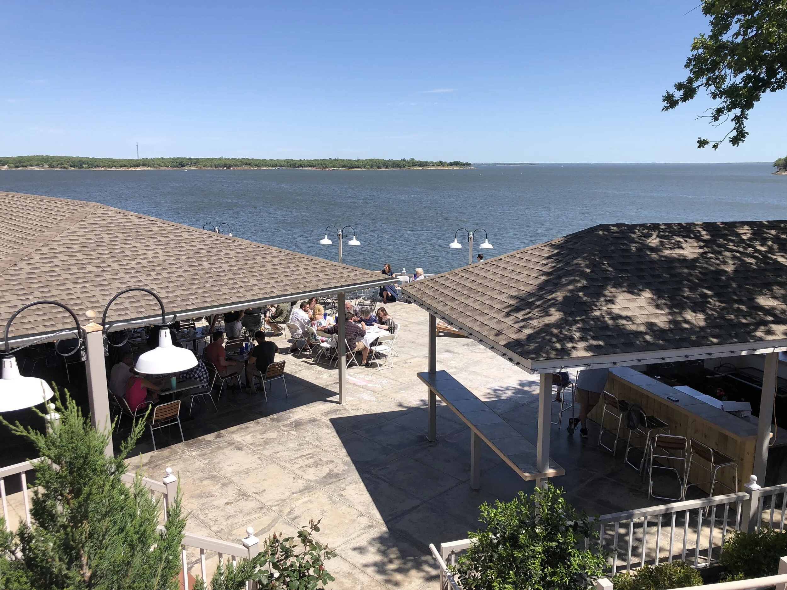 Outdoor dining area by a lake with people seated at tables, two small structures with tiled roofs, and a view of the water in the background.