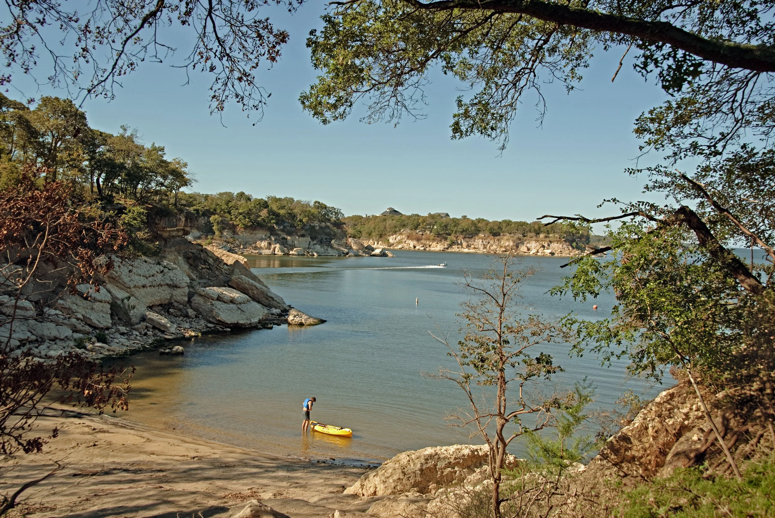 Person preparing a yellow kayak on the beach near a calm body of water, with rocky cliffs and trees in the background on a clear day.