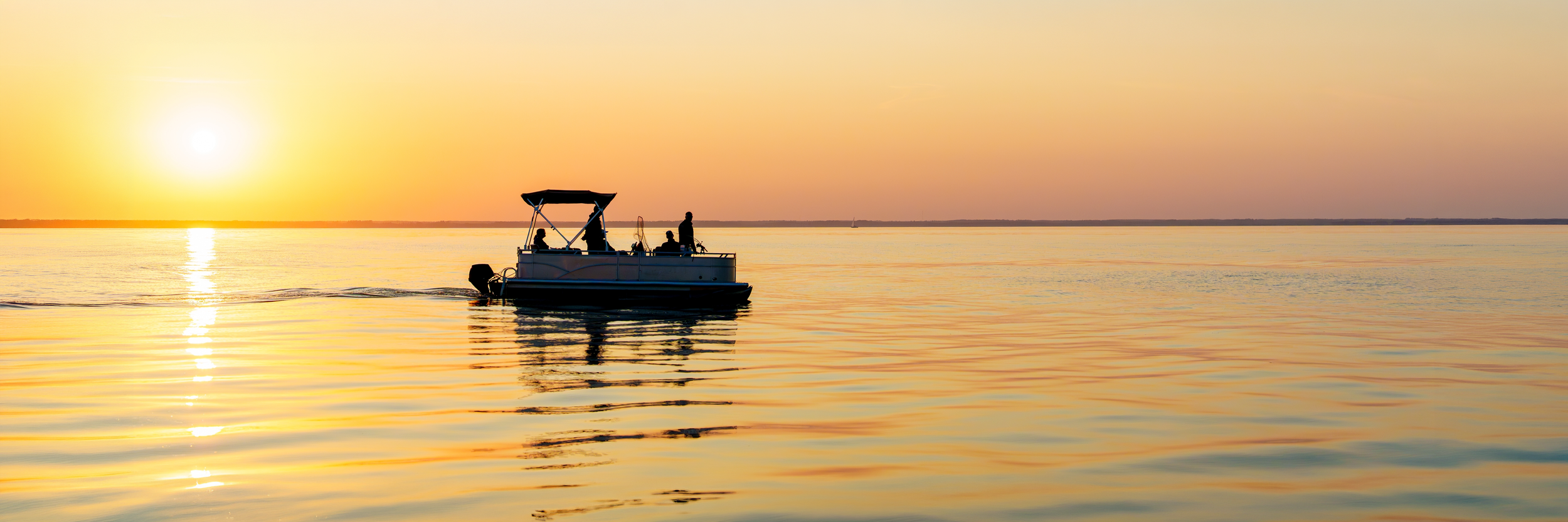 A boat with three people on board sailing on calm water during sunset or sunrise, with a clear sky and the sun near the horizon.