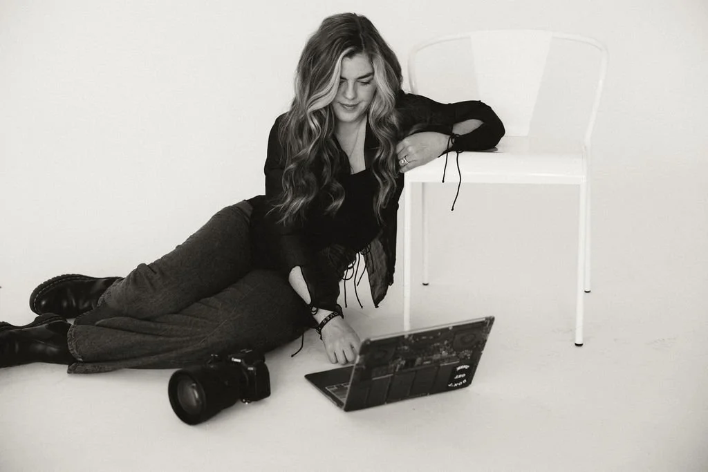 A young woman with wavy hair lying on the floor beside a chair, looking at a laptop. A camera is placed nearby on the floor.