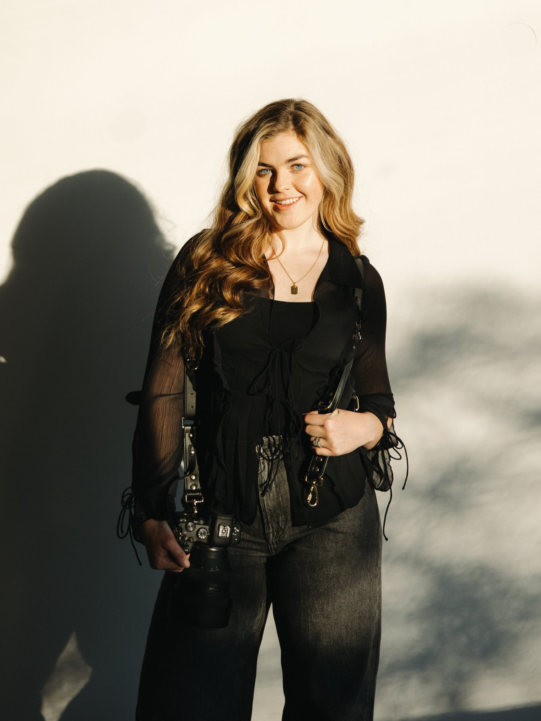 Young woman with wavy blonde hair smiling, holding a camera, dressed in black with a camera strap and a gold necklace, standing against a plain light background with a shadow behind her.
