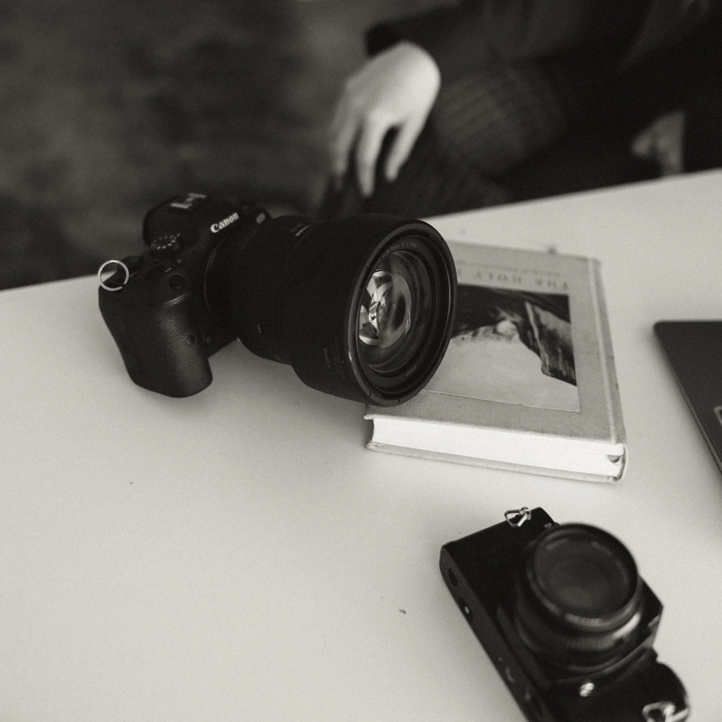 A black Canon camera with a zoom lens sitting on a white surface beneath a dark shelf, with a person reaching for it.