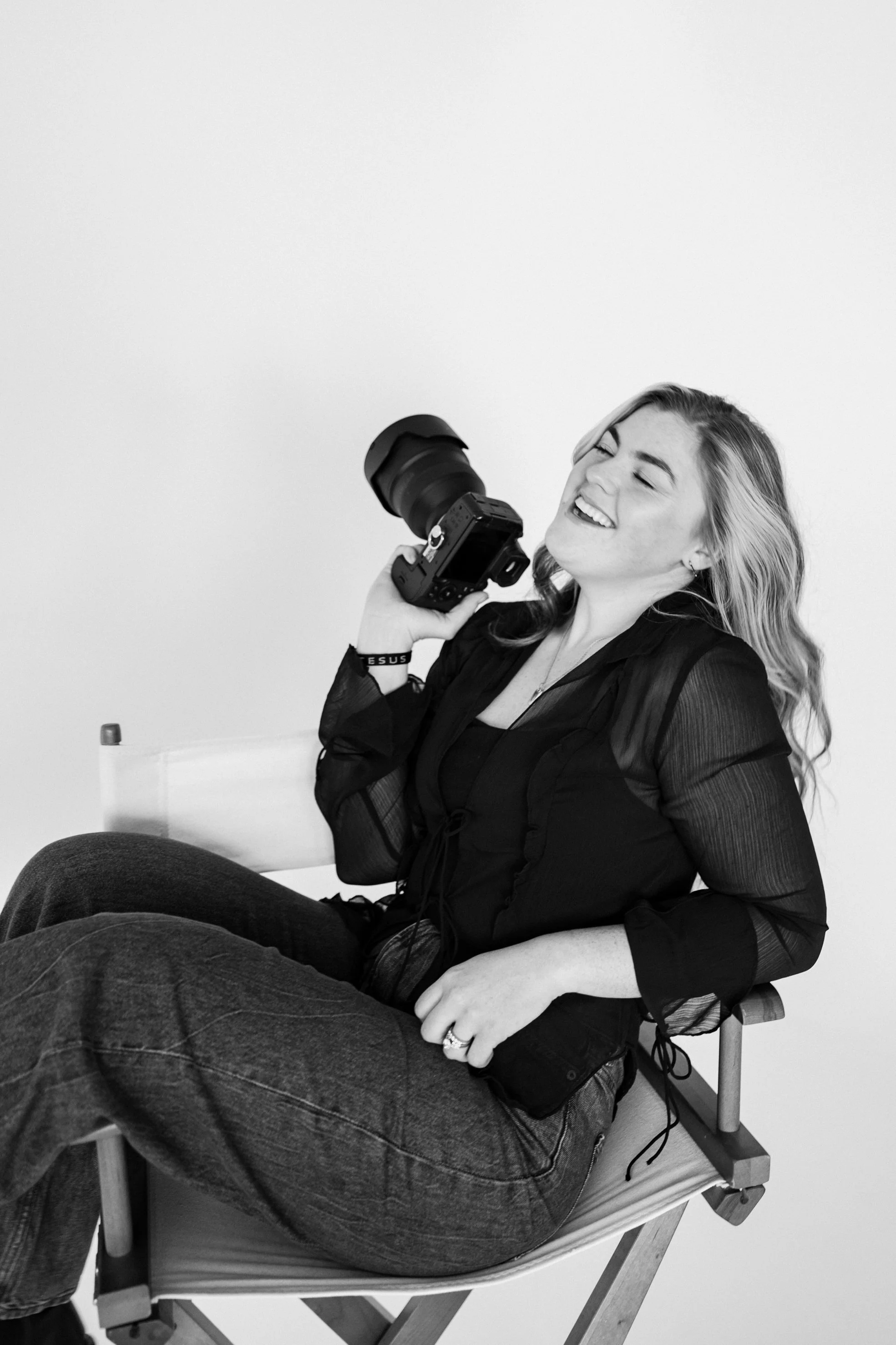 A woman with long hair, smiling and winking, sitting on a wooden chair, holding a camera with a large lens, wearing a dark top and jeans, against a plain background.