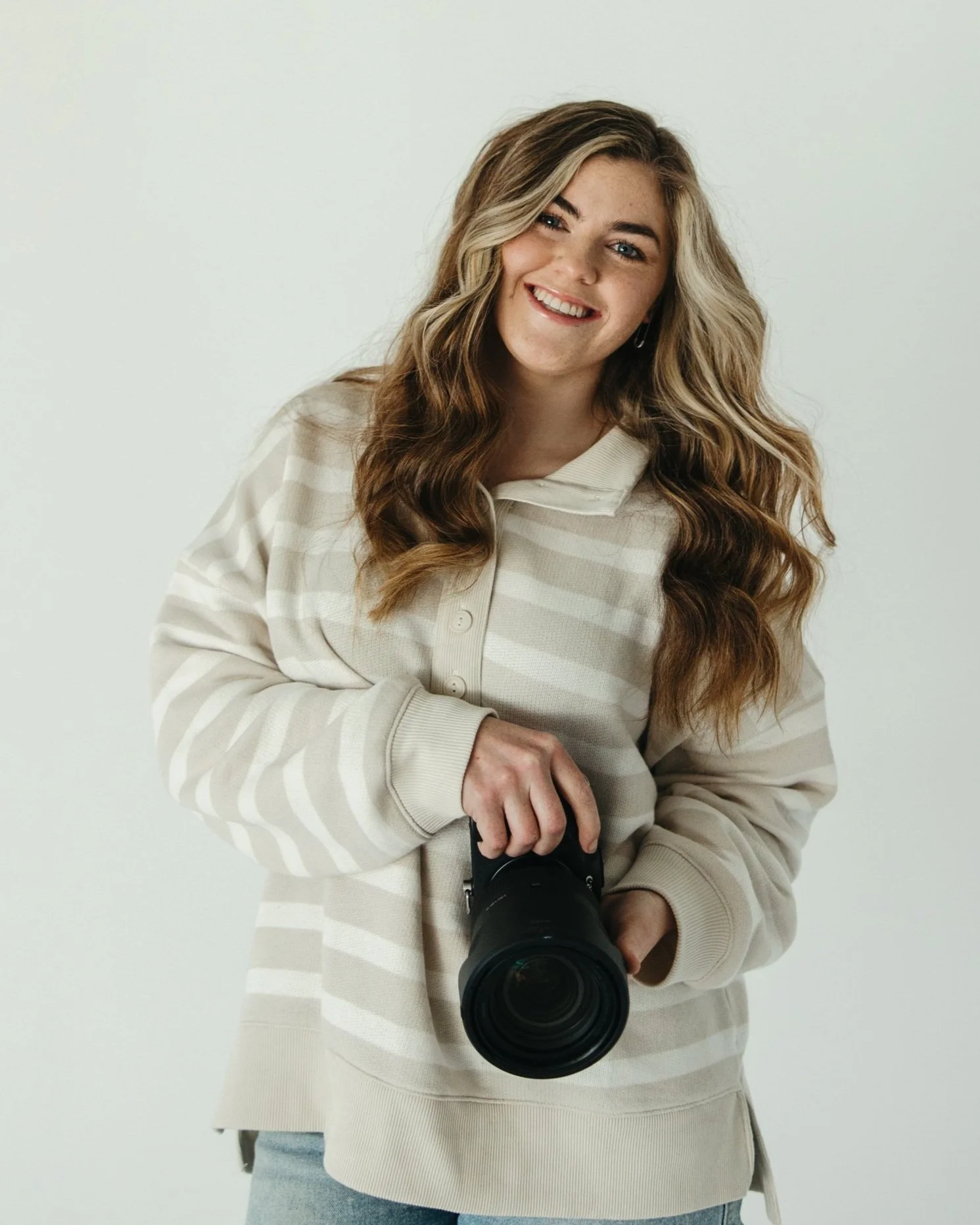 A young woman with long wavy hair, blue eyes, and a bright smile, holding a professional camera. She is wearing a cream and beige striped hoodie and stands against a plain white background.