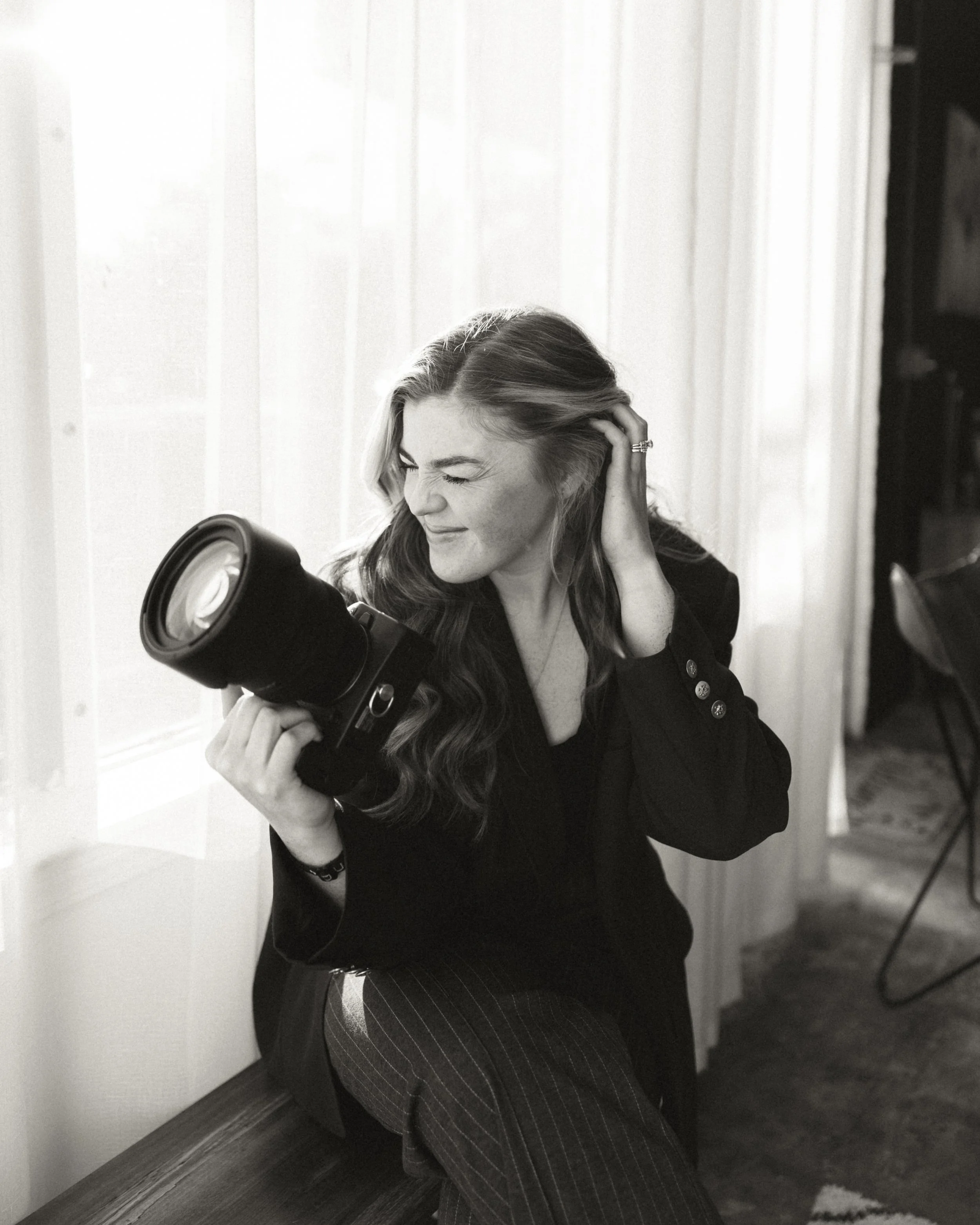 A woman with wavy hair smiling, wearing a black top, holding hair with one hand, and looking downward. She is seated, with a corner of a room with white vertical panels and a piece of black furniture or curtain in the background.