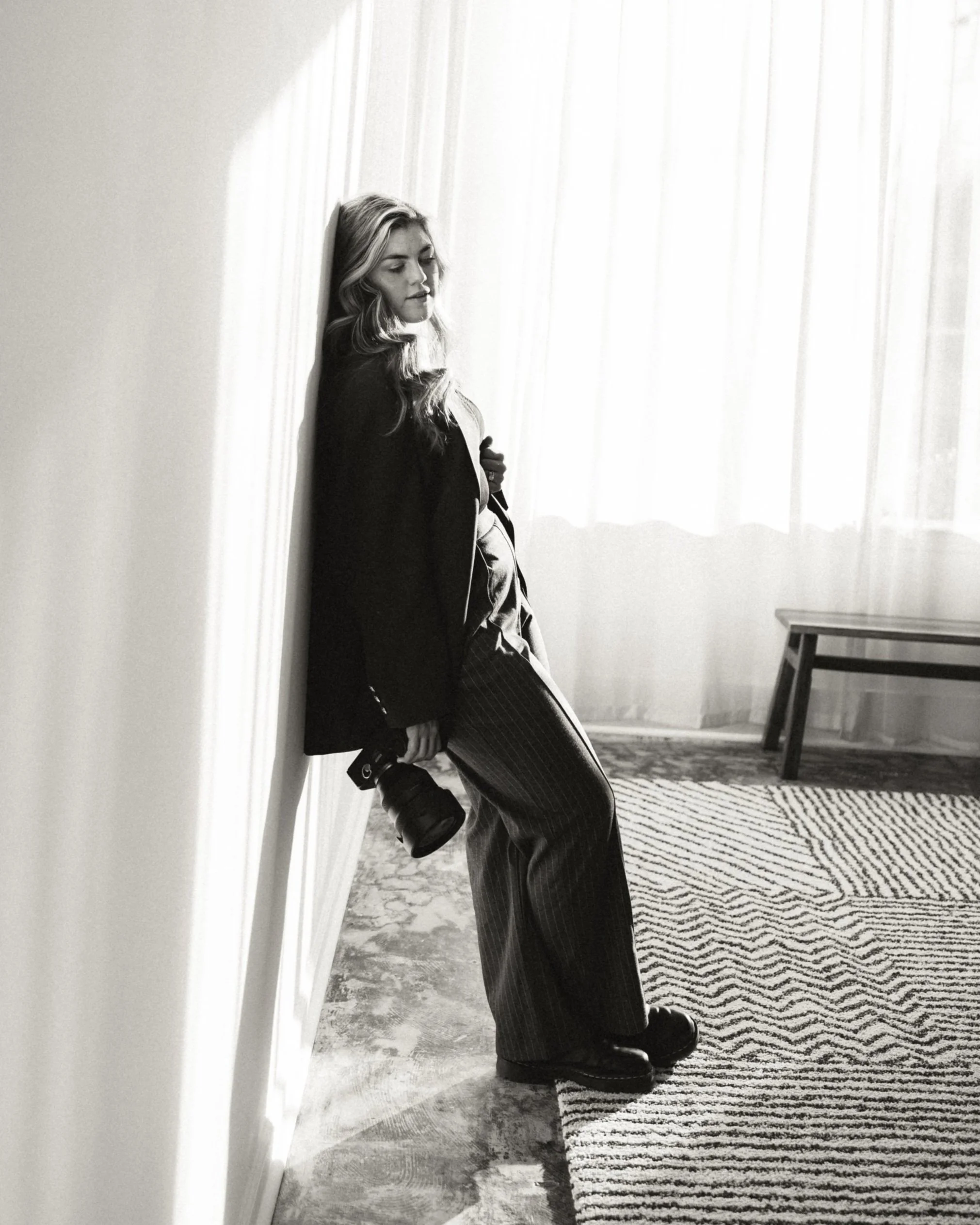 Black and white photo of a woman leaning against a wall, holding a camera, with a patterned rug and furniture in the background.