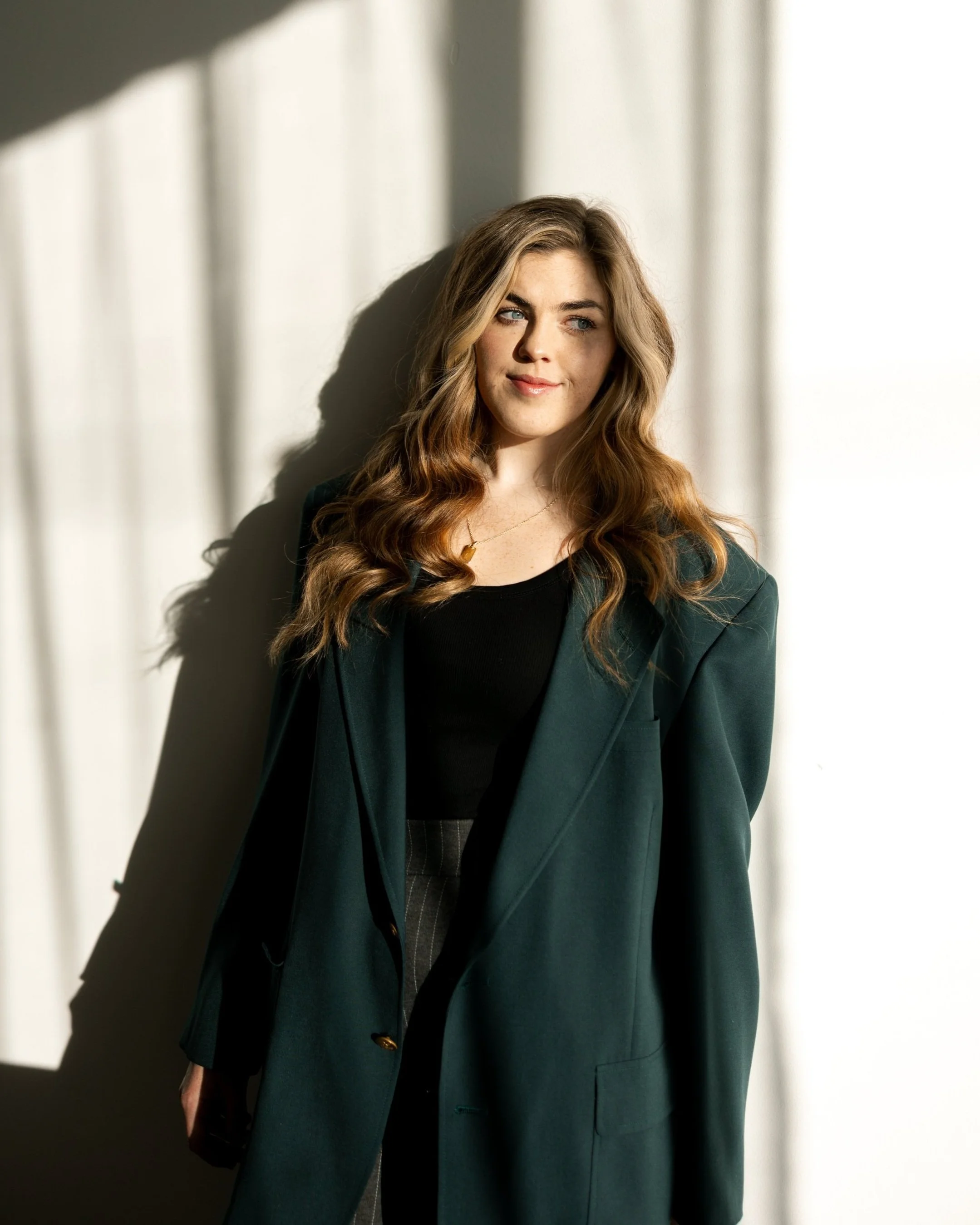 A woman with long wavy light brown hair stands near a wall casting a shadow, looking slightly to the side with a subtle smile, wearing a dark green blazer and black top, with a gold heart necklace.