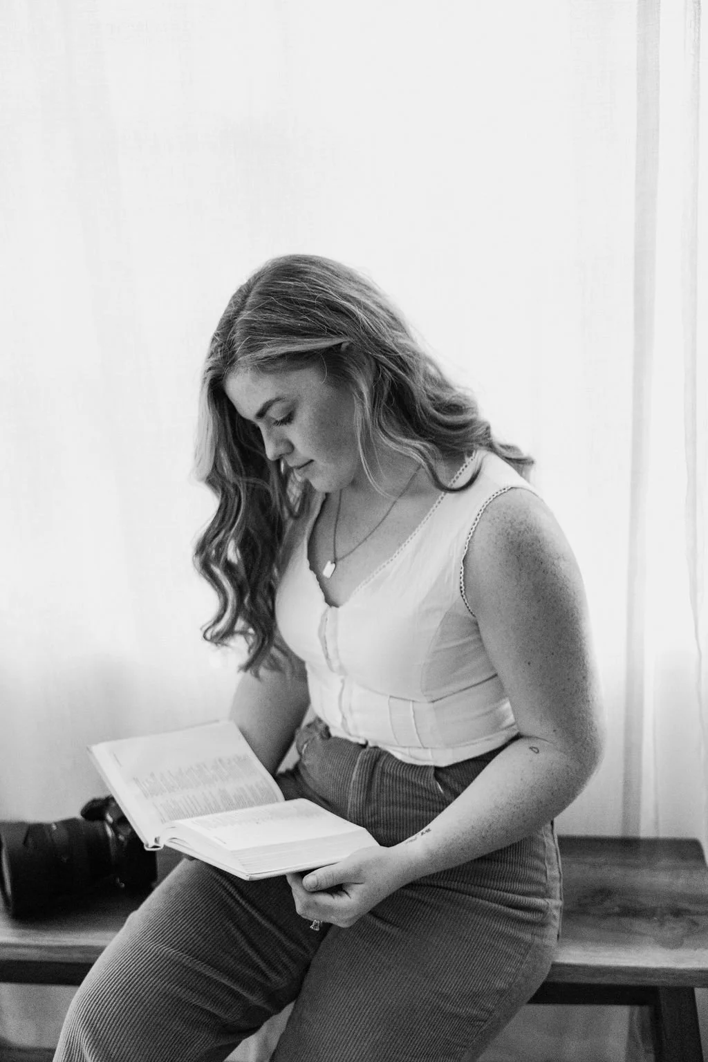 A woman with long wavy hair sitting on a bench, reading a book, wearing a sleeveless top and high-waisted pants, in black and white photograph.