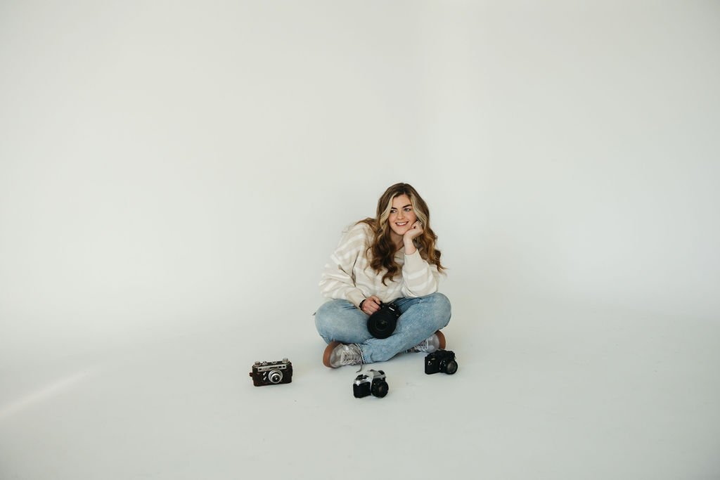 A young woman with long wavy hair sitting cross-legged on the floor, smiling, surrounded by vintage and modern cameras against a plain light background.
