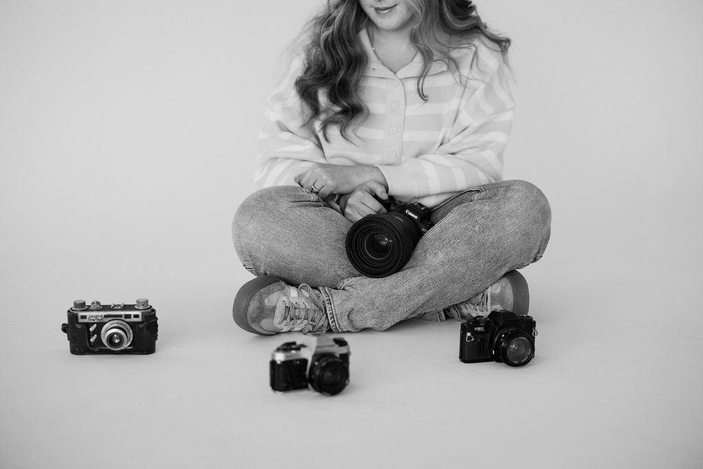 A woman with long hair sitting cross-legged on the floor, surrounded by vintage and modern cameras, holding a camera in her lap.