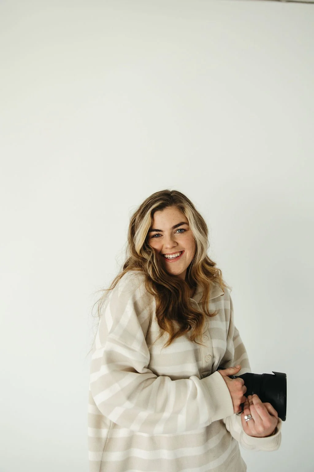 A woman with wavy blonde hair smiling and holding a camera, wearing a beige striped sweatshirt, against a plain light background.