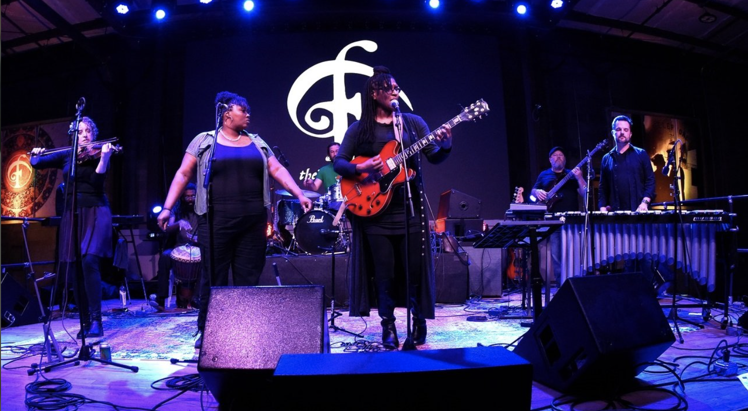 Musicians performing on stage, with a large logo in the background. The group includes a violinist, a singer with a tambourine, a guitarist, a bassist, and a keyboardist.