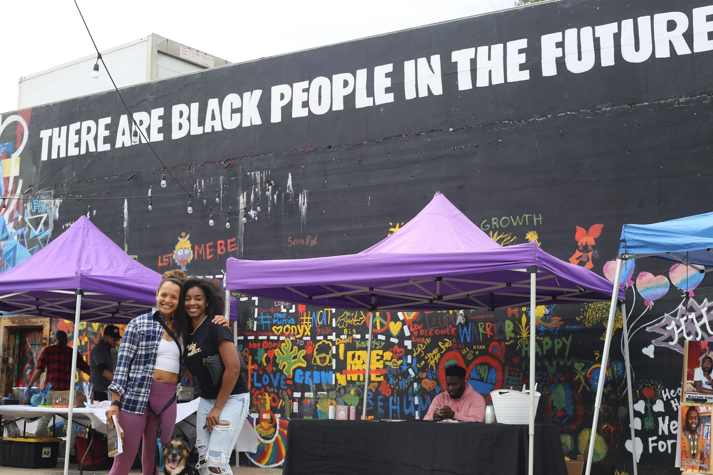 Two women smiling and standing in front of a vibrant mural wall with black background, featuring colorful graffiti art and a large message that says 'THERE ARE BLACK PEOPLE IN THE FUTURE.' They are at the Black Business Expo.