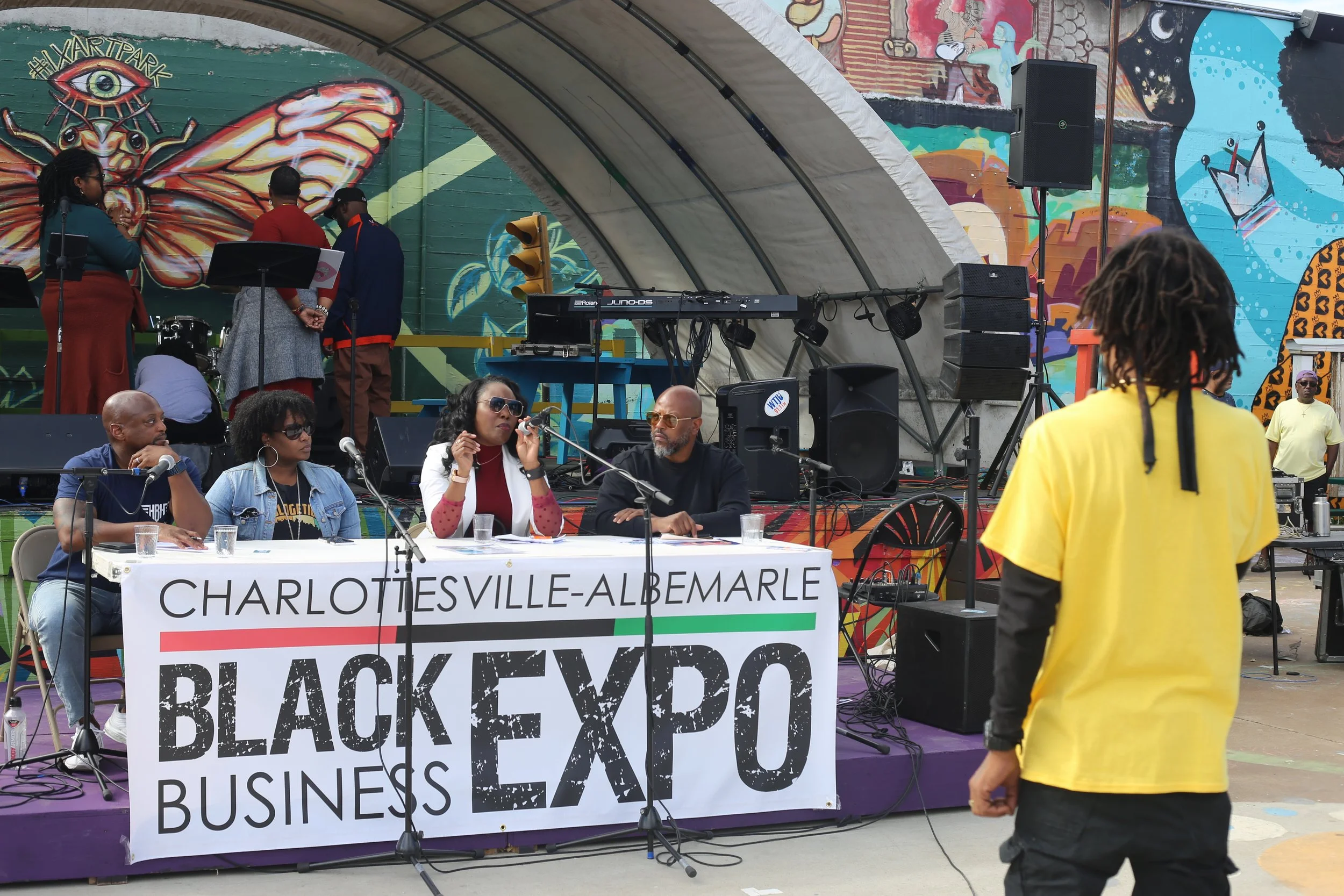 A panel of five people seated at a table with a banner reading 'Charlottesville-Albemarle Black Expo Business' during an outdoor event. Behind them is a colorful mural and a stage with musical equipment. A man with dreadlocks wearing a yellow shirt stands in the foreground, facing the panel.