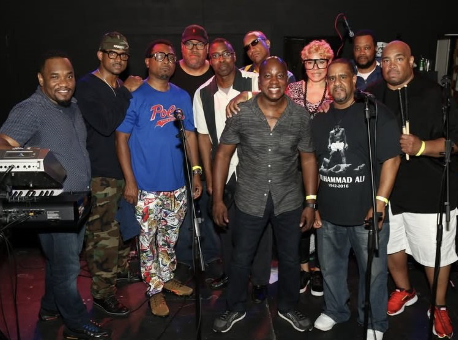 Group of diverse people on stage with musical equipment, smiling for a group photo during a performance or event.