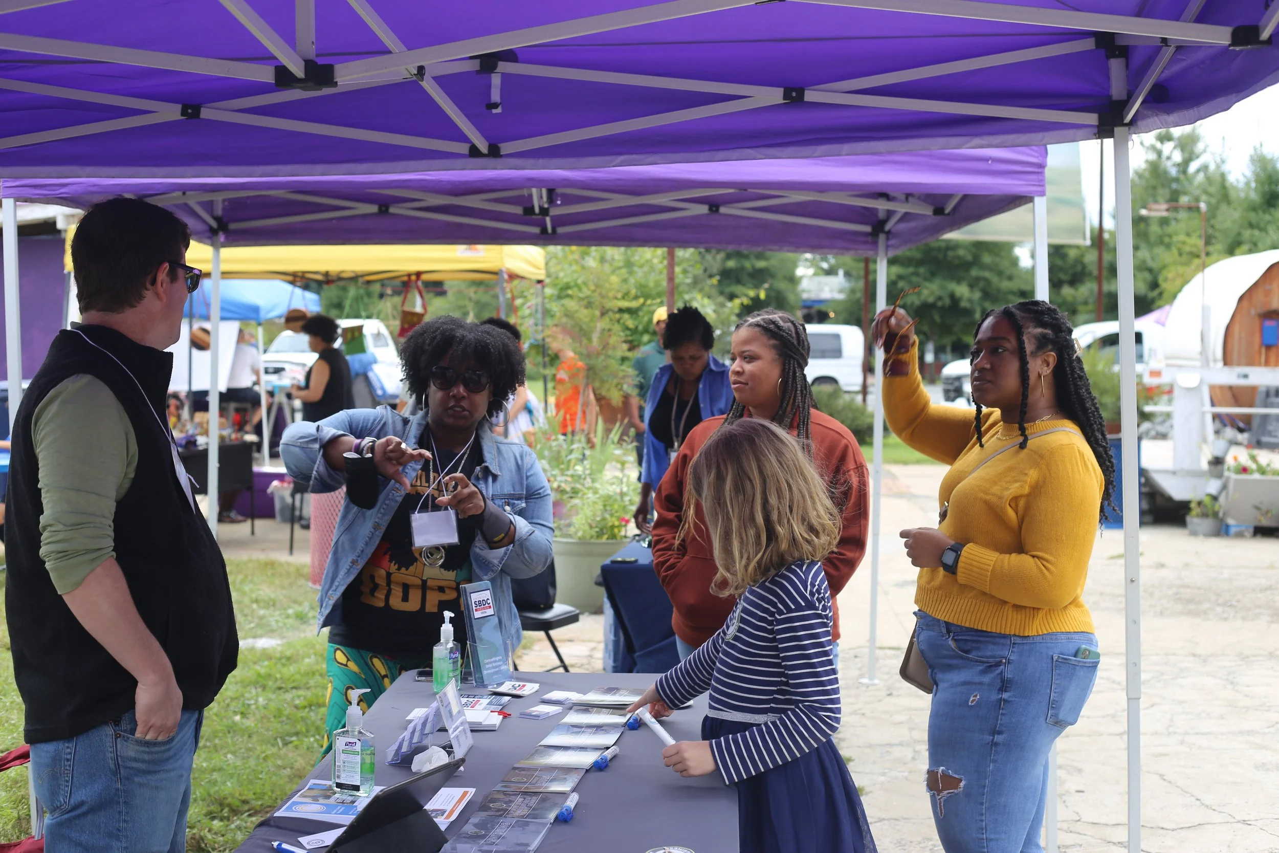 People gathered around a table at an outdoor fair booth under a purple canopy, engaging in conversation, with other vendor booths and vehicles in the background.