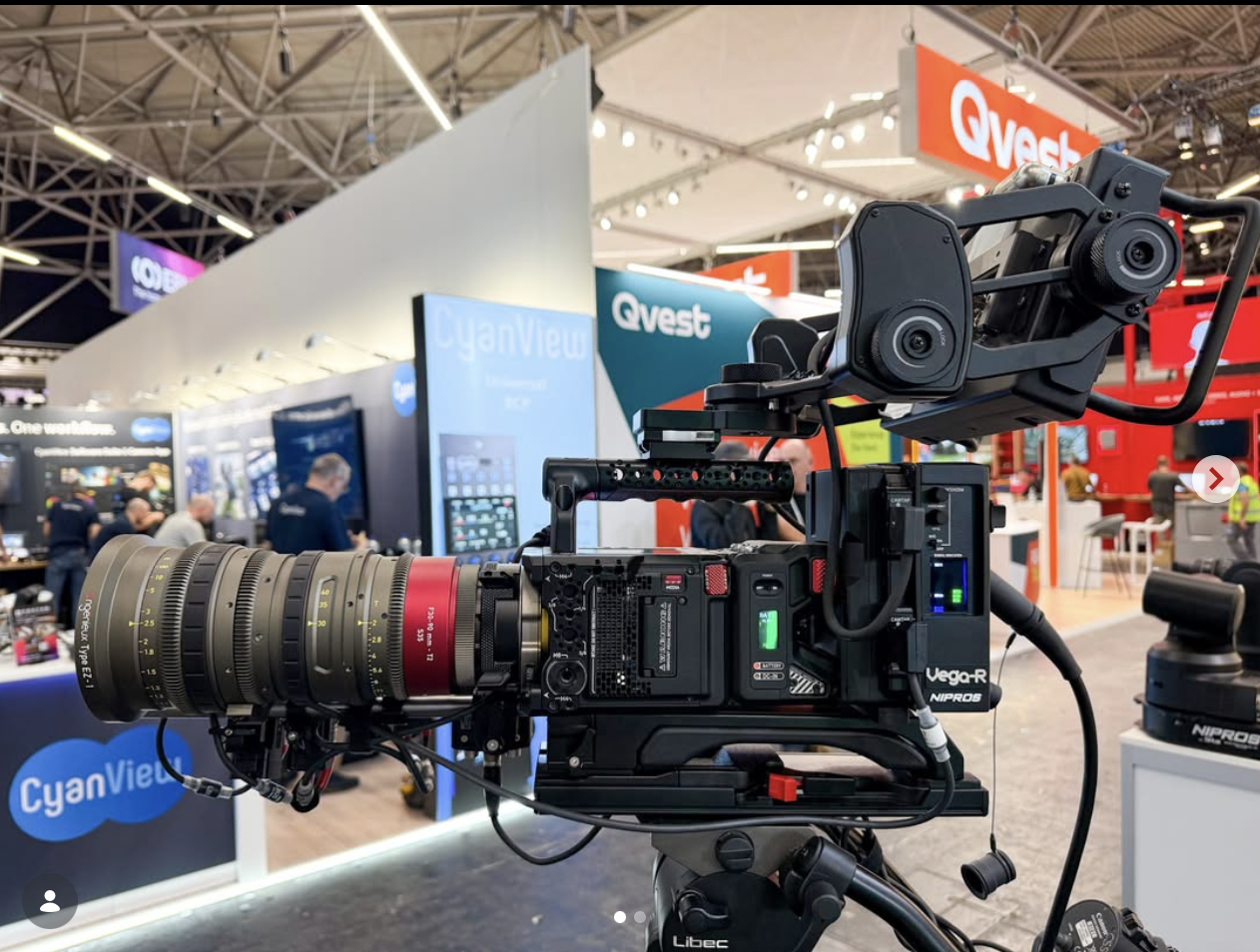 Professional video camera with a large zoom lens mounted on a tripod, set up indoors at a tech convention or trade show, with exhibition booths and people in the background.