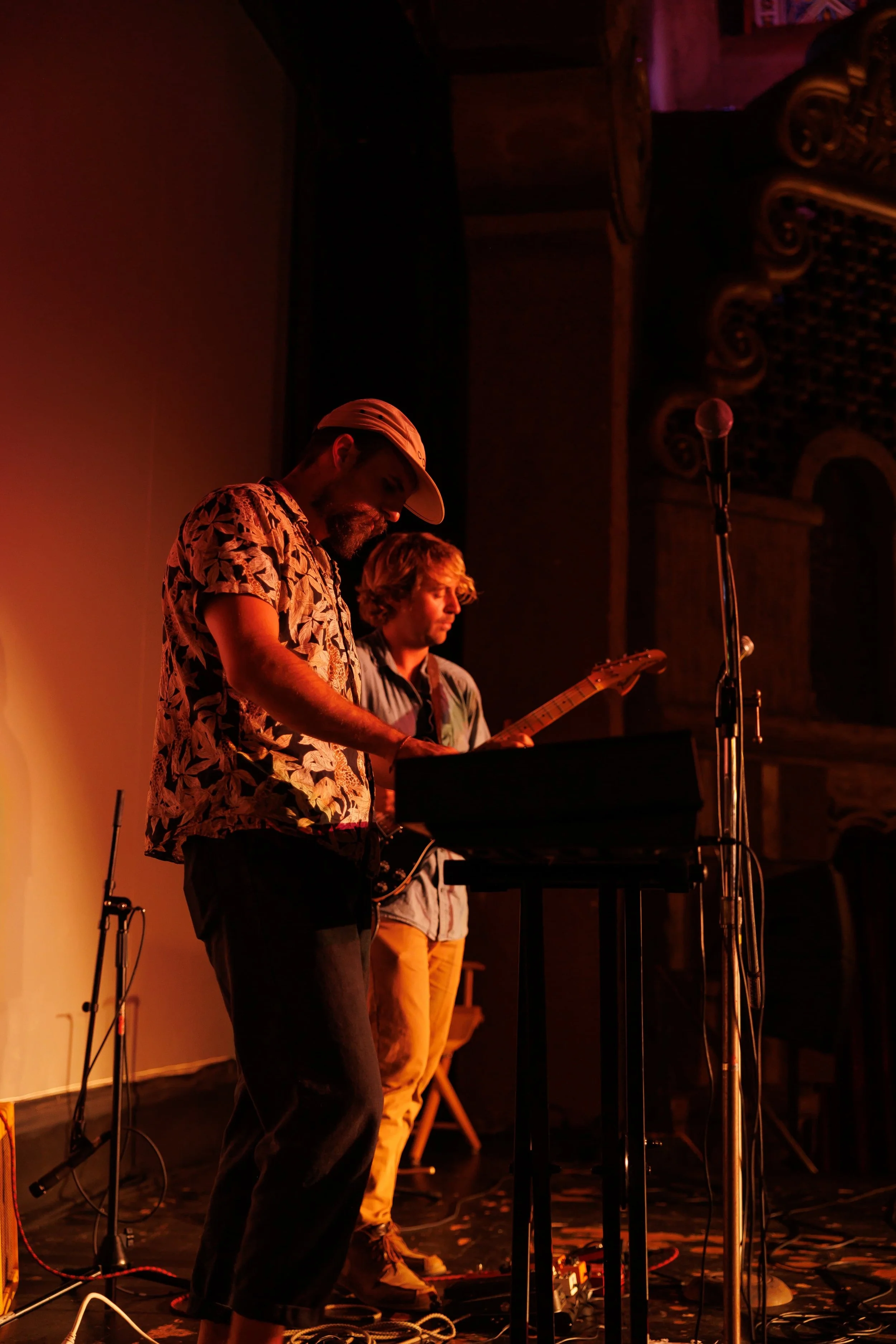 Two musicians performing on stage, one playing keyboard and the other playing guitar, with warm stage lighting and an ornate background.