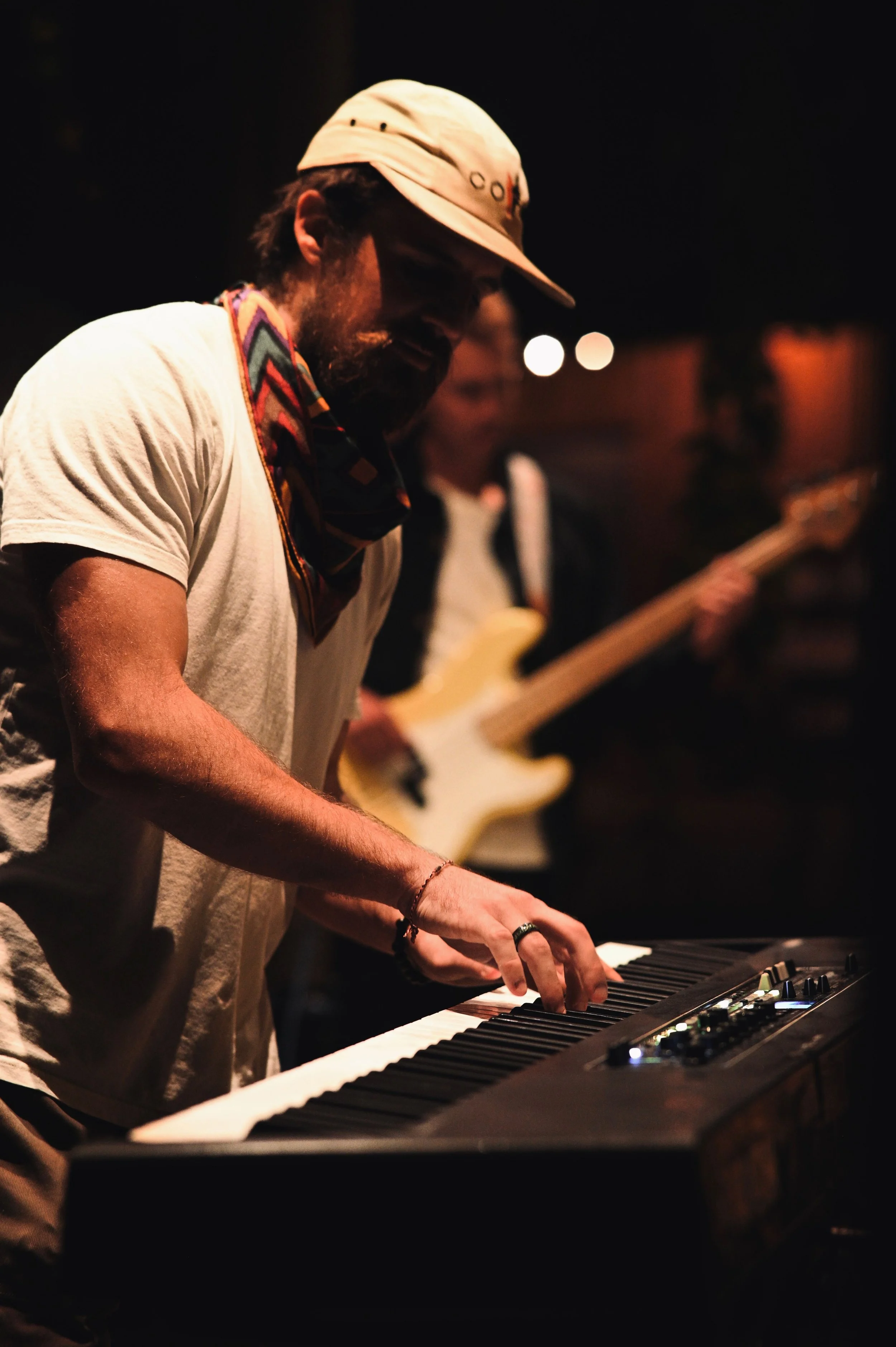 A man playing a keyboard with a bassist in the background in a dimly lit setting.