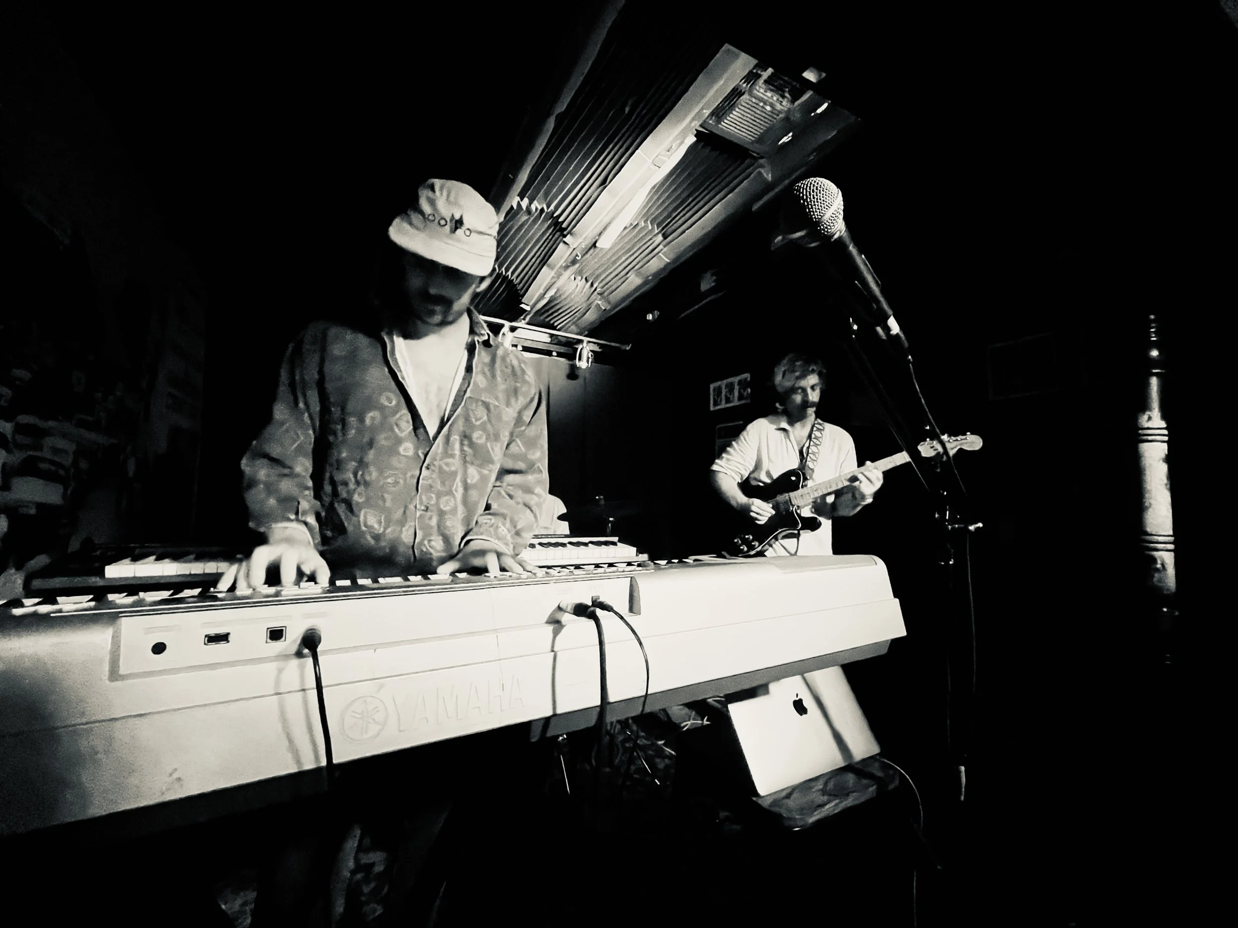 Two musicians performing on stage: one playing a keyboard and the other playing an electric guitar, in a dimly lit venue.