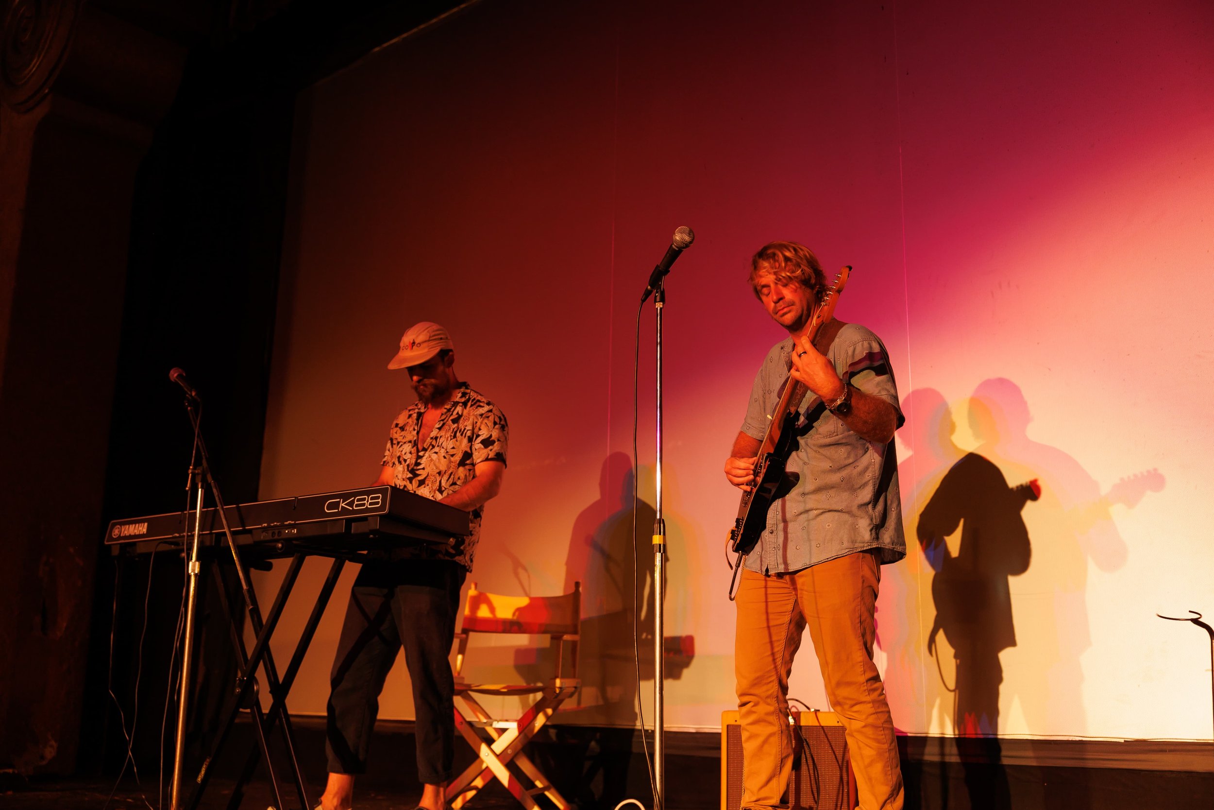 Two musicians performing on stage, one playing keyboard and the other playing guitar, with colorful lighting creating shadows on the wall behind them.
