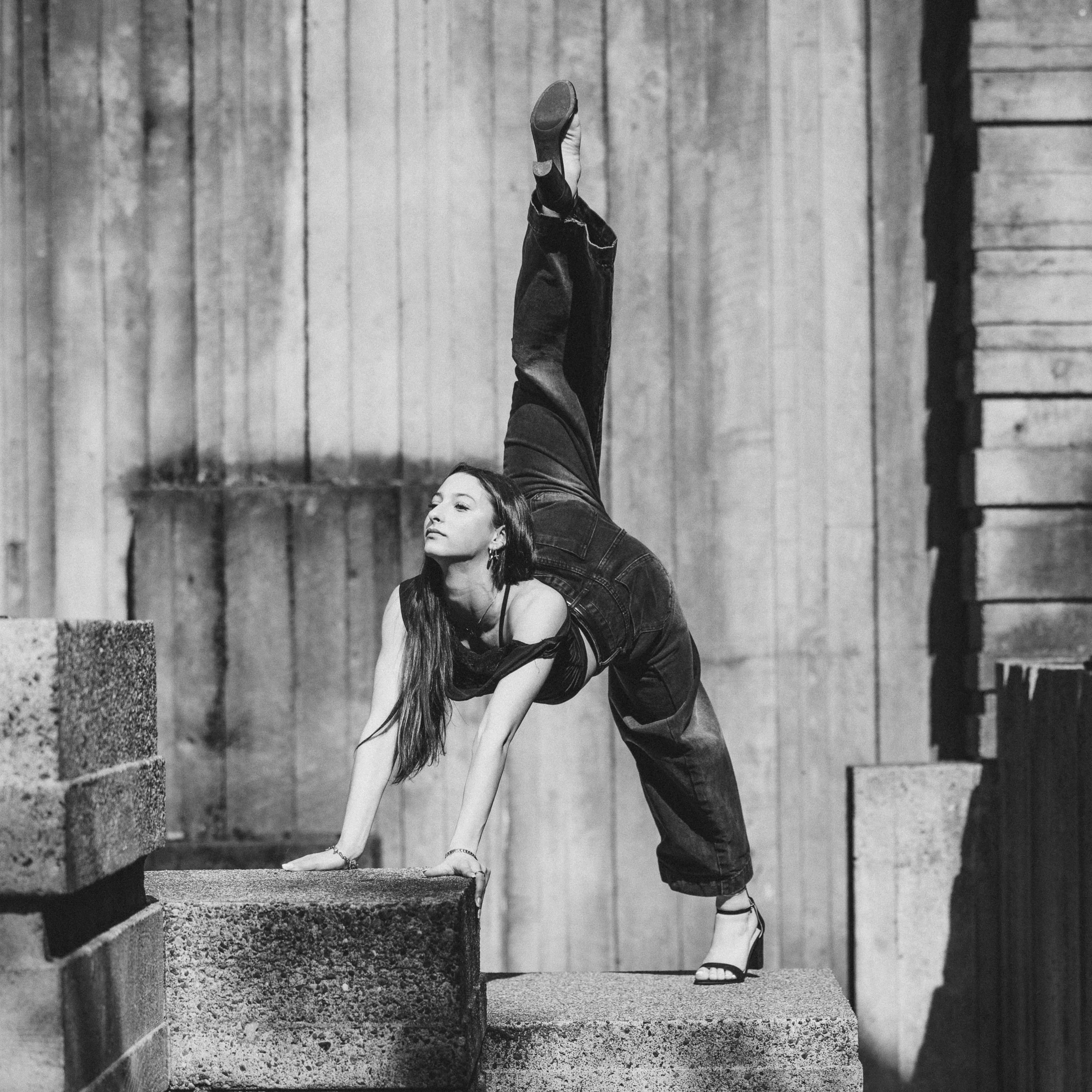 A girl performing a dance pose with two arms on the floor and leg in a penché on concrete blocks outdoors. Captured in black and white.