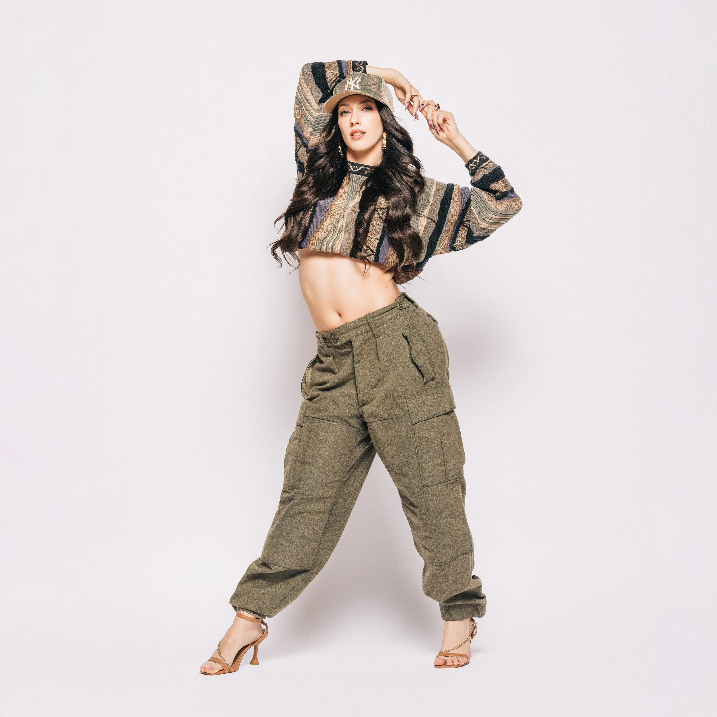 Young woman with long dark hair posing in trendy streetwear, wearing a crop top, loose cargo pants, high heels, and a cap, against a plain white background.
