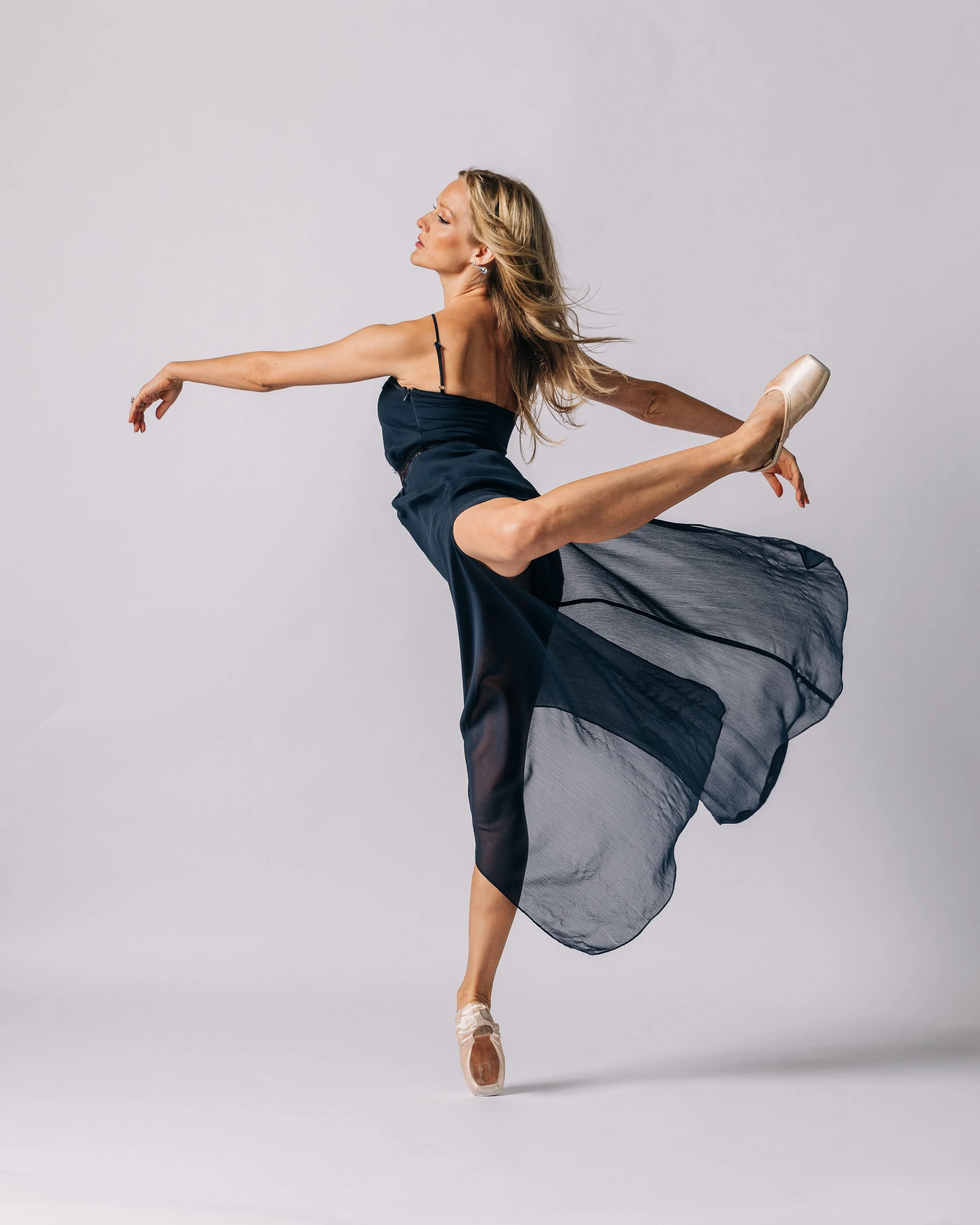 Ballet dancer in a pose, wearing a black dress and pointe shoes, with one leg extended behind her and arms outstretched against a plain background.