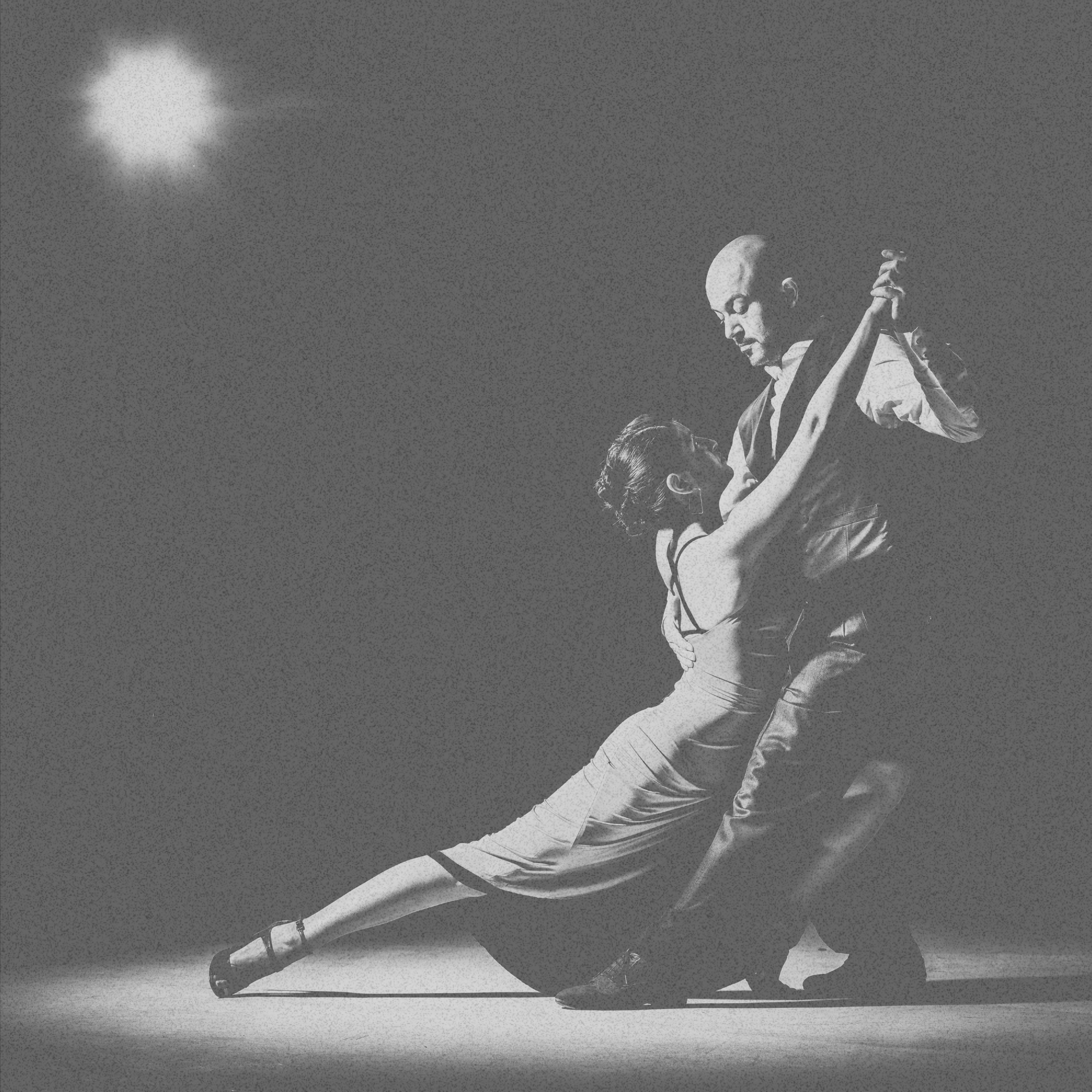 A black and white photo of a man and woman dancing. The woman is wearing a dress and high heels, extending her leg backwards while the man, dressed in formal attire, holds her close as they dance under a spotlight.