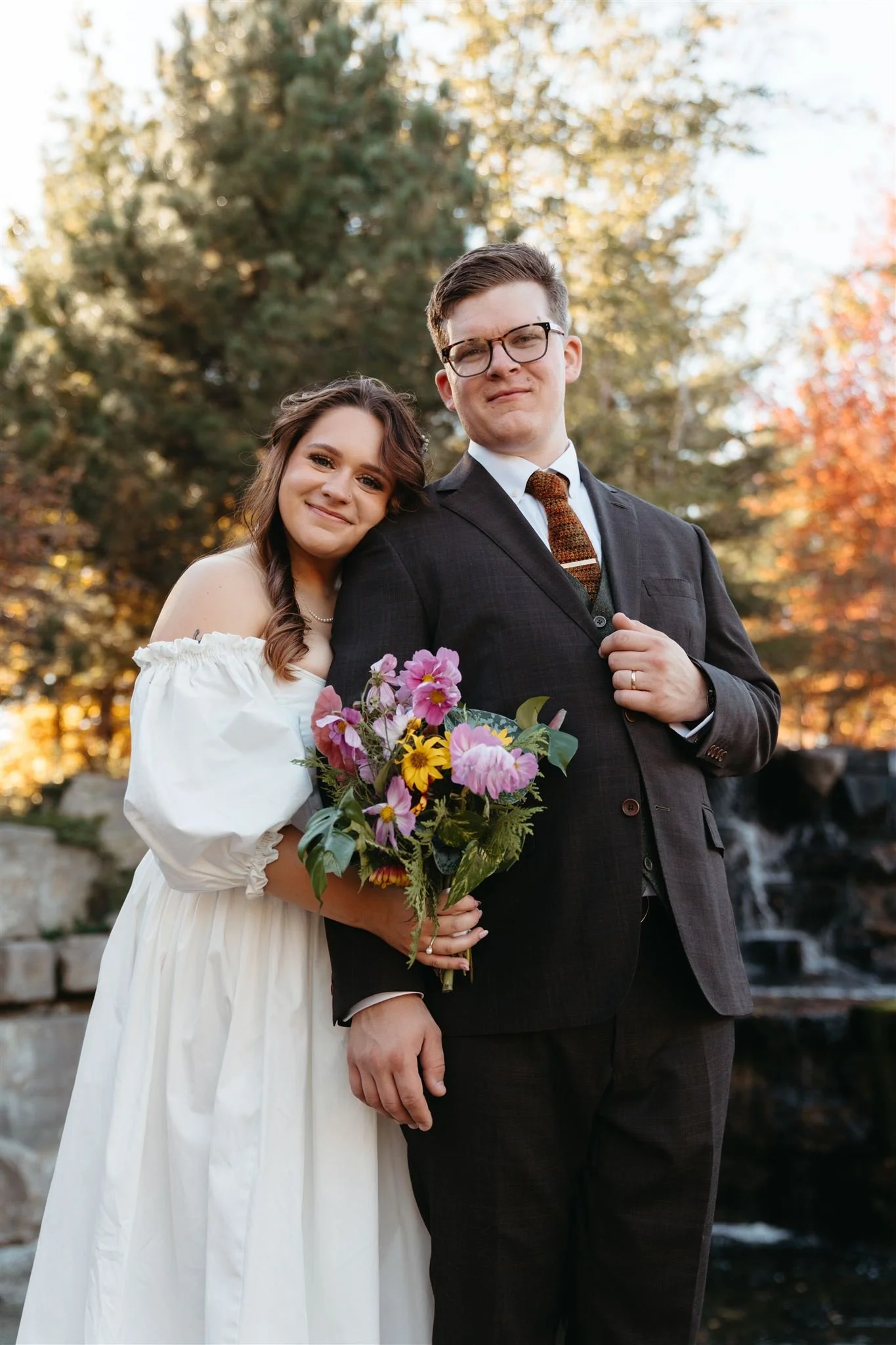 A couple when outdoor for a wedding, the woman in a white dress holding a bouquet of flowers, leaning her head on the man in a suit, with trees and a waterfall in the background.