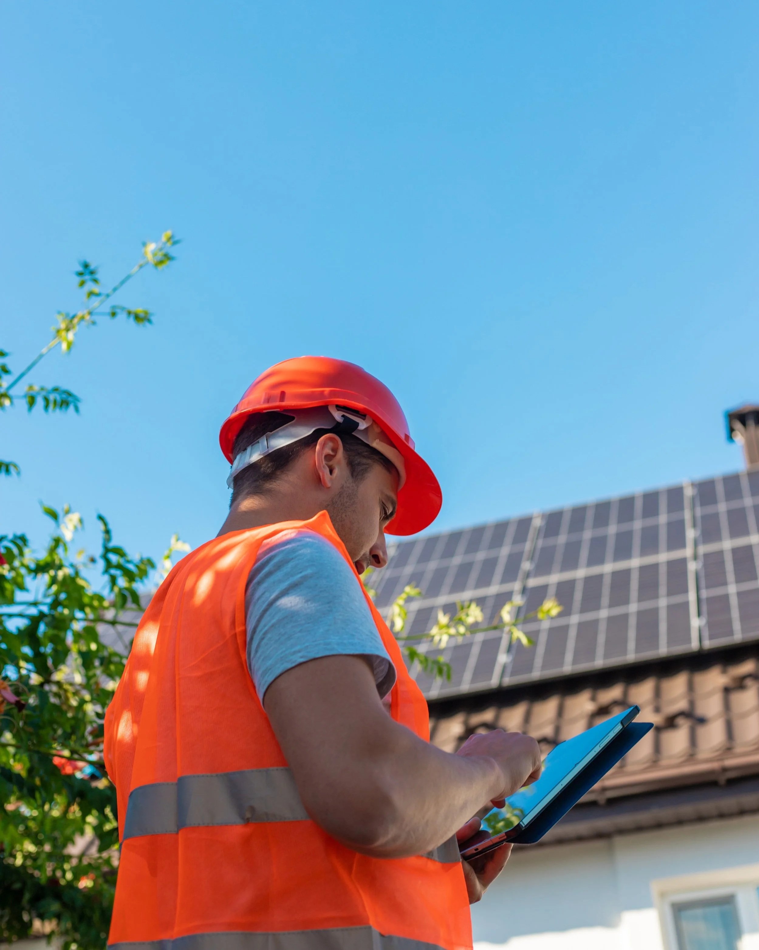 A construction worker wearing a red safety helmet and orange safety vest looks at a tablet outside near a house with solar panels on the roof under a blue sky.