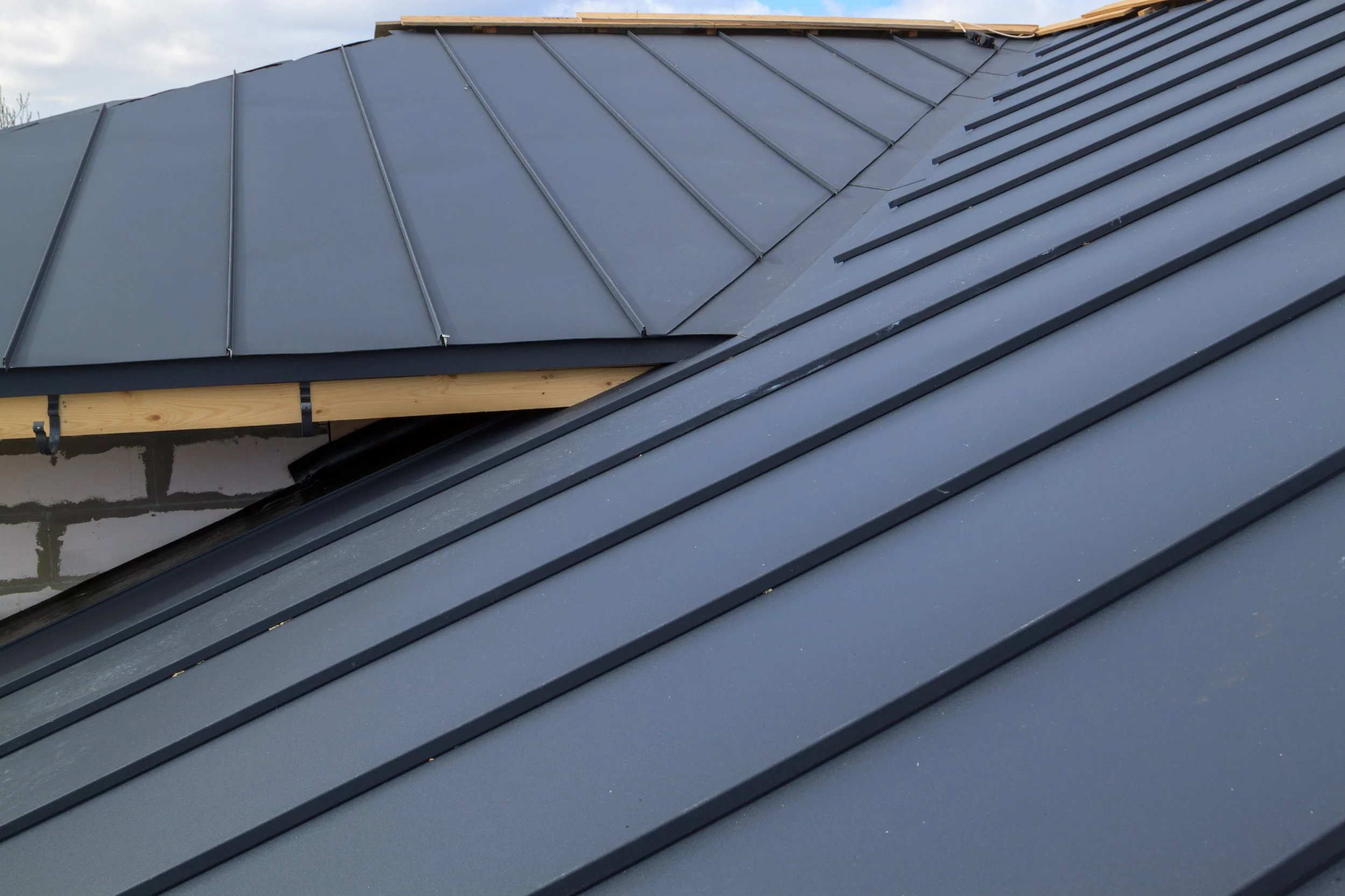 Close-up of a metal roof with a new black finish, showing the panels and ridges, with part of a roof section in the background, under a cloudy sky.