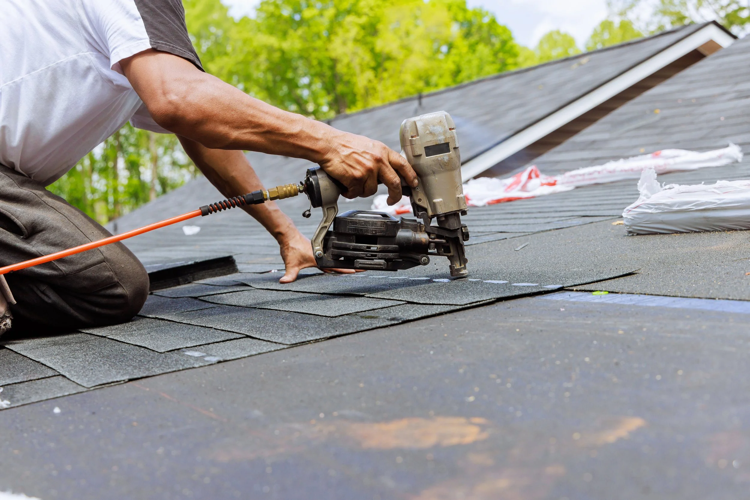 Person installing or repairing asphalt shingles on a roof using a pneumatic nail gun during daytime.