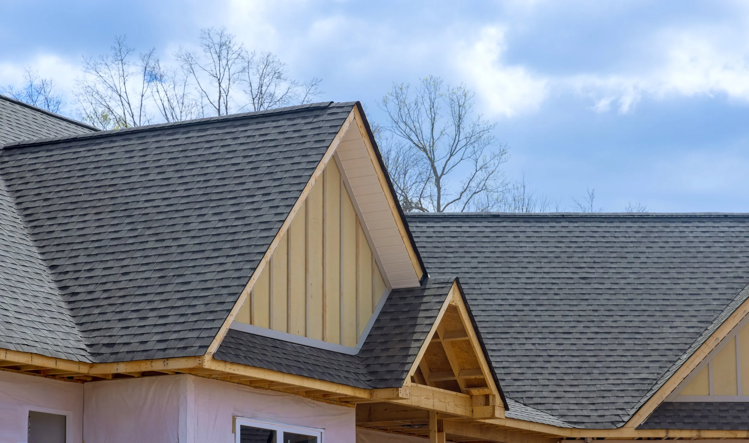 Close-up of a house roof with dark gray shingles and under construction gables with yellow sheathing and exposed wooden framing, against a background of leafless trees and a partly cloudy sky.
