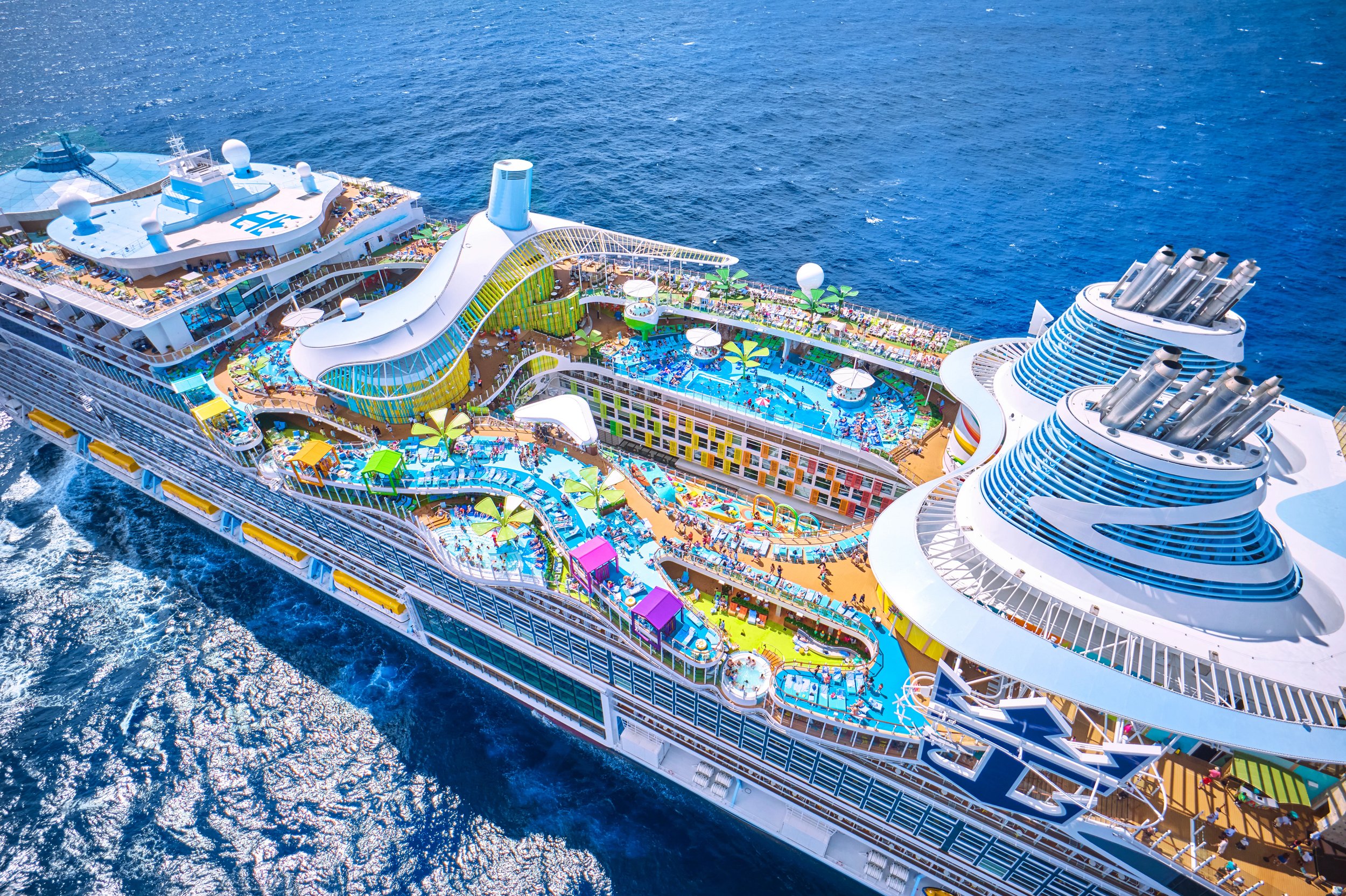 An aerial view of Icon of the seas, a large cruise ship, with multiple pools, water slides, and a sunbathing area, sailing in the open ocean.