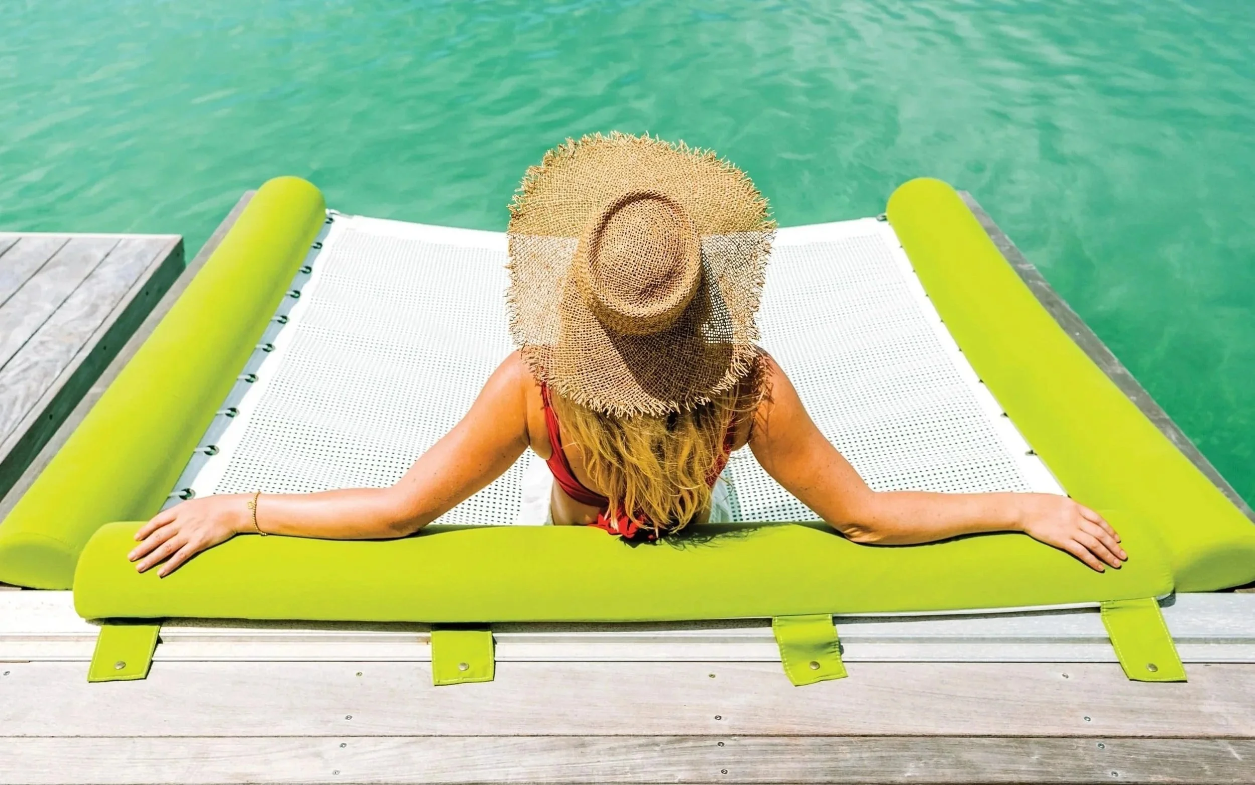 Woman relaxing on a floating lounger on a clear blue-green body of water, wearing a straw hat and a red swimsuit.