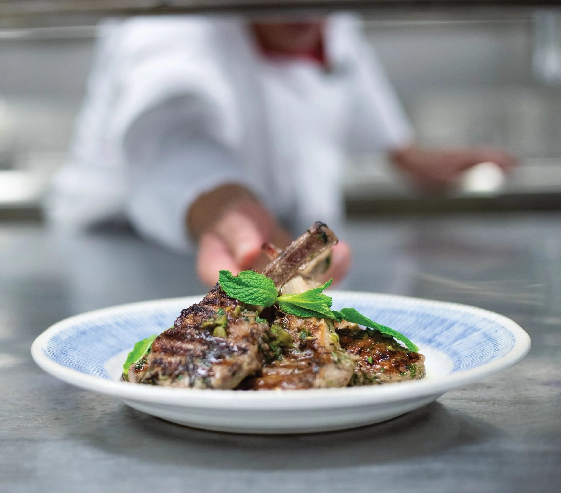 Close-up of a plate with grilled lamb chops garnished with mint leaves at Chops on Royal Caribbean Cruise ship, in a restaurant kitchen with a chef in the background.