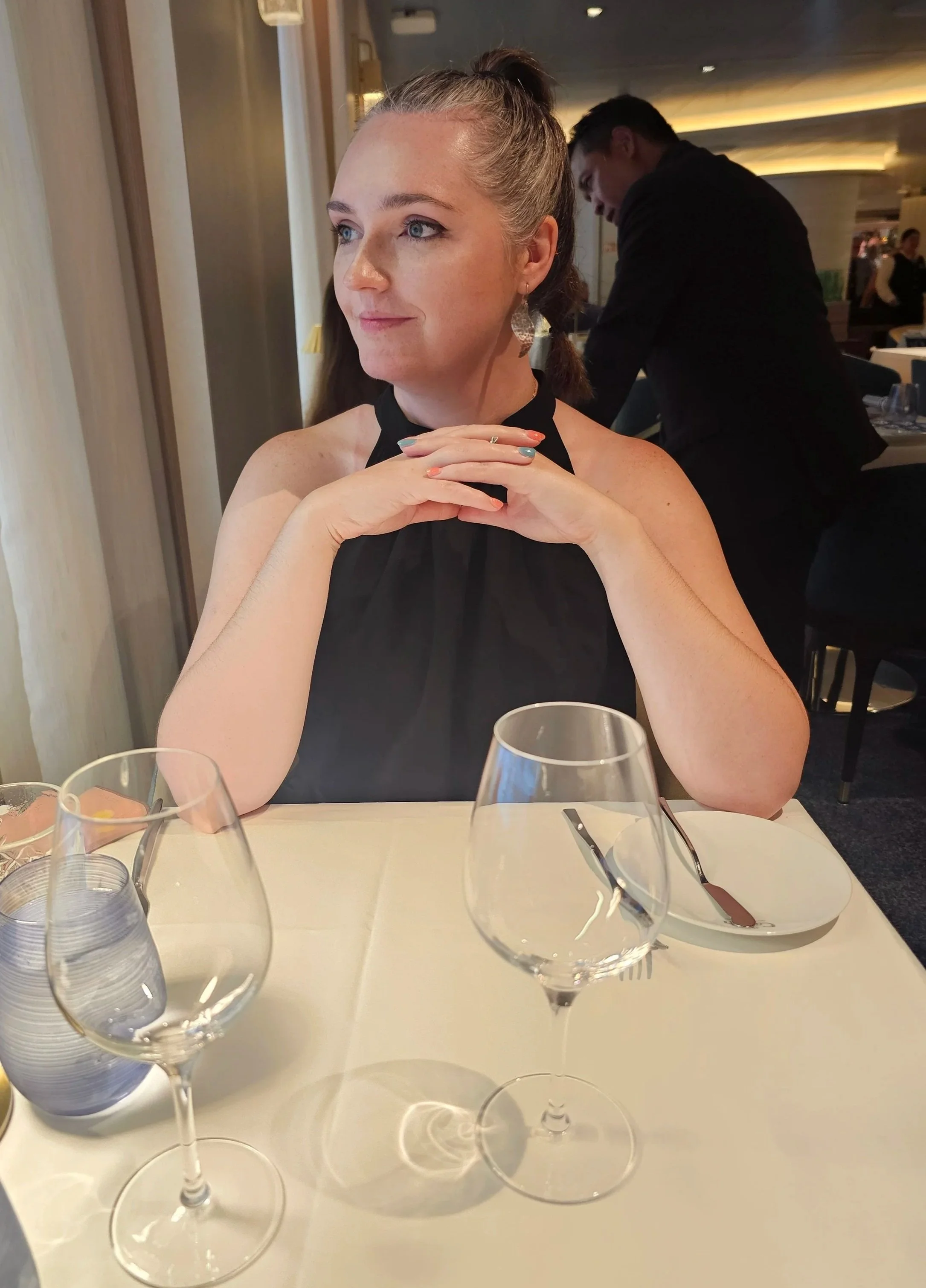 A woman with light skin and blonde hair tied back, sitting at a restaurant table with empty wine glasses, a plate, and water glasses. She is wearing a black sleeveless top and silver earrings, and looks thoughtful while resting her chin on her hands.