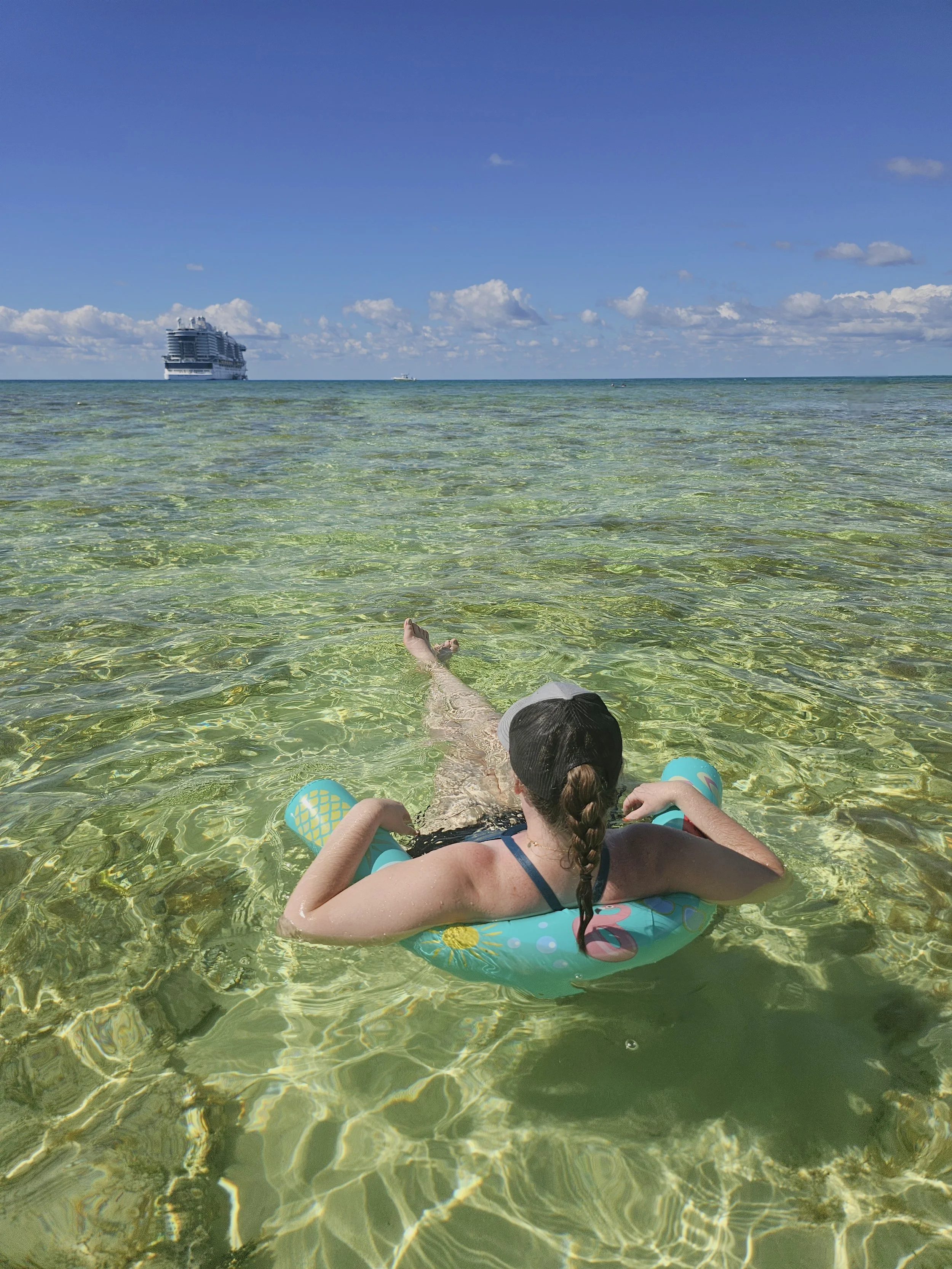 A girl with a braid, wearing a black swimsuit, a cap, and floaties, relaxing on an inflatable ring in clear ocean water with a cruise ship in the distance under a blue sky with clouds.