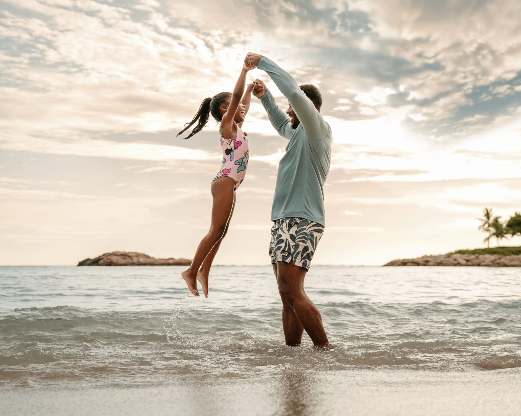A man and a young girl playing and laughing in the shallow ocean water during sunset at Aulani resort in Hawaii, with the girl jumping into the water and the man holding her hands.