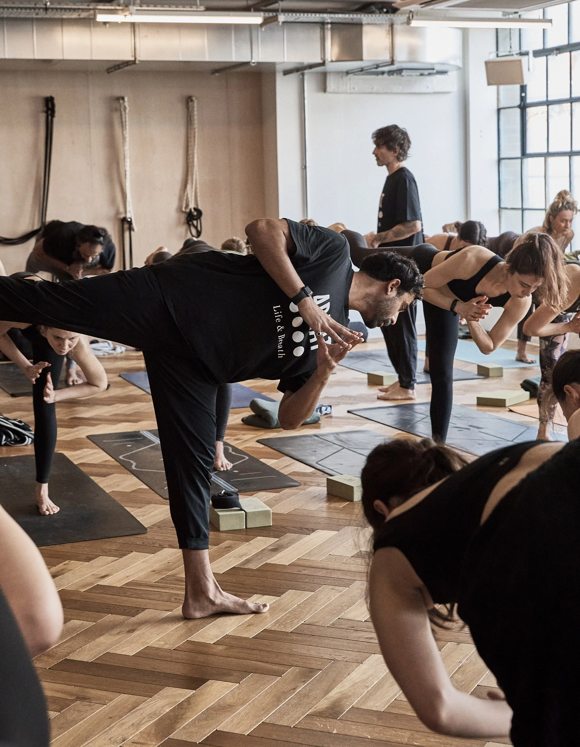 People practicing yoga in a studio, some in downward dog pose, with yoga mats and blocks, in a well-lit room with large windows.