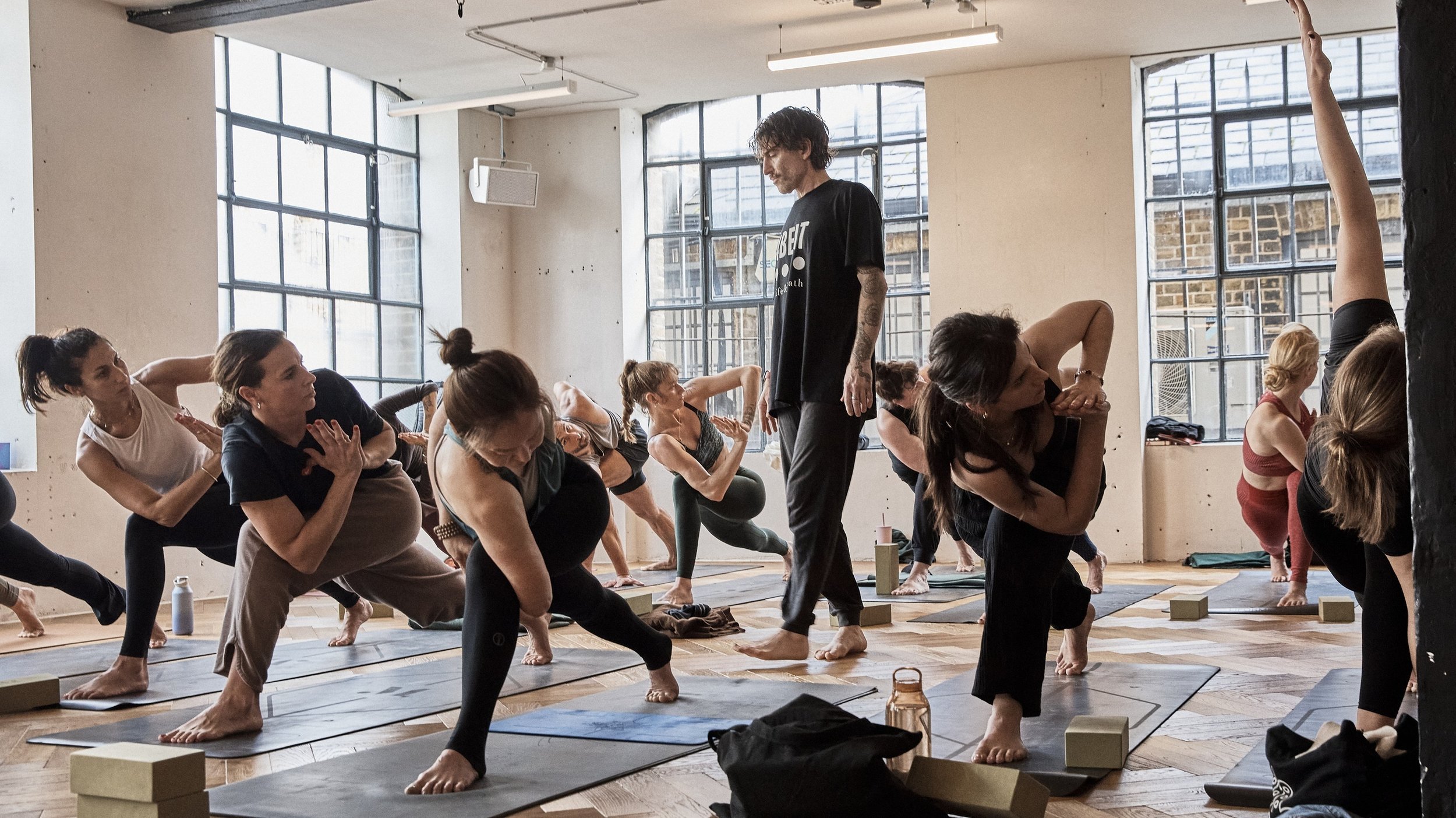 Group of people doing yoga in a studio with large windows and wooden floors, guided by an instructor.