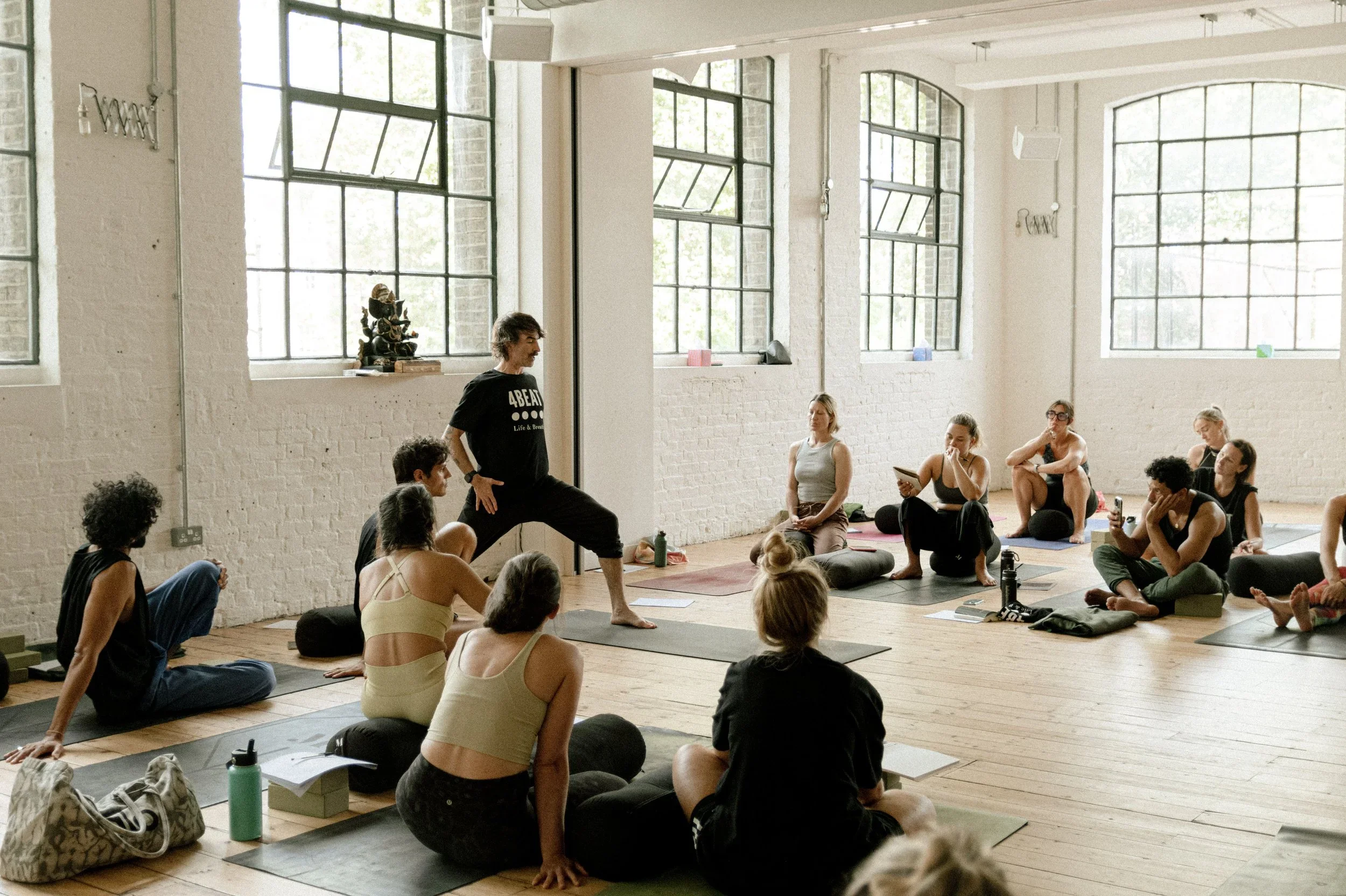 A group fitness class in a bright studio with large windows and white brick walls. Participants are sitting on yoga mats, listening to the instructor in a black T-shirt demonstrating a stretch.