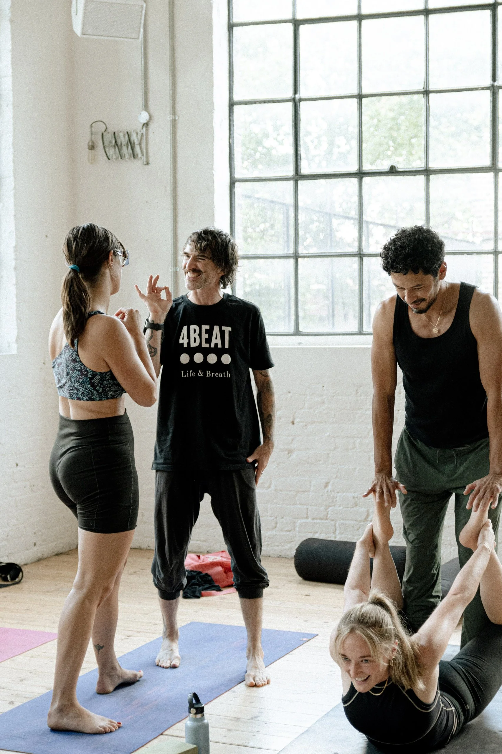 Group of people participating in a yoga or fitness class indoors, with a woman lying on her stomach with her arms raised, two instructors assisting, and one talking to another participant.