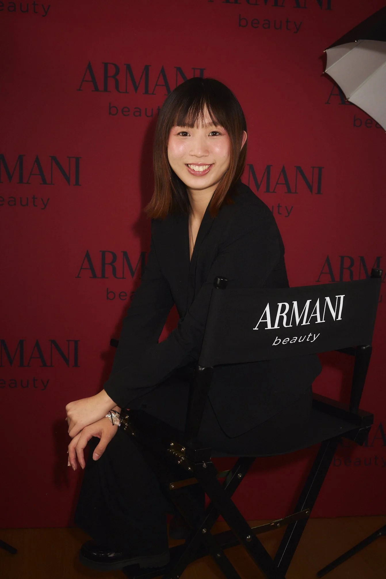 A woman sitting on a black Armani Beauty chair in front of a red backdrop with 'Armani beauty' printed repeatedly. She has shoulder-length brown hair, a smile, and is dressed in black.
