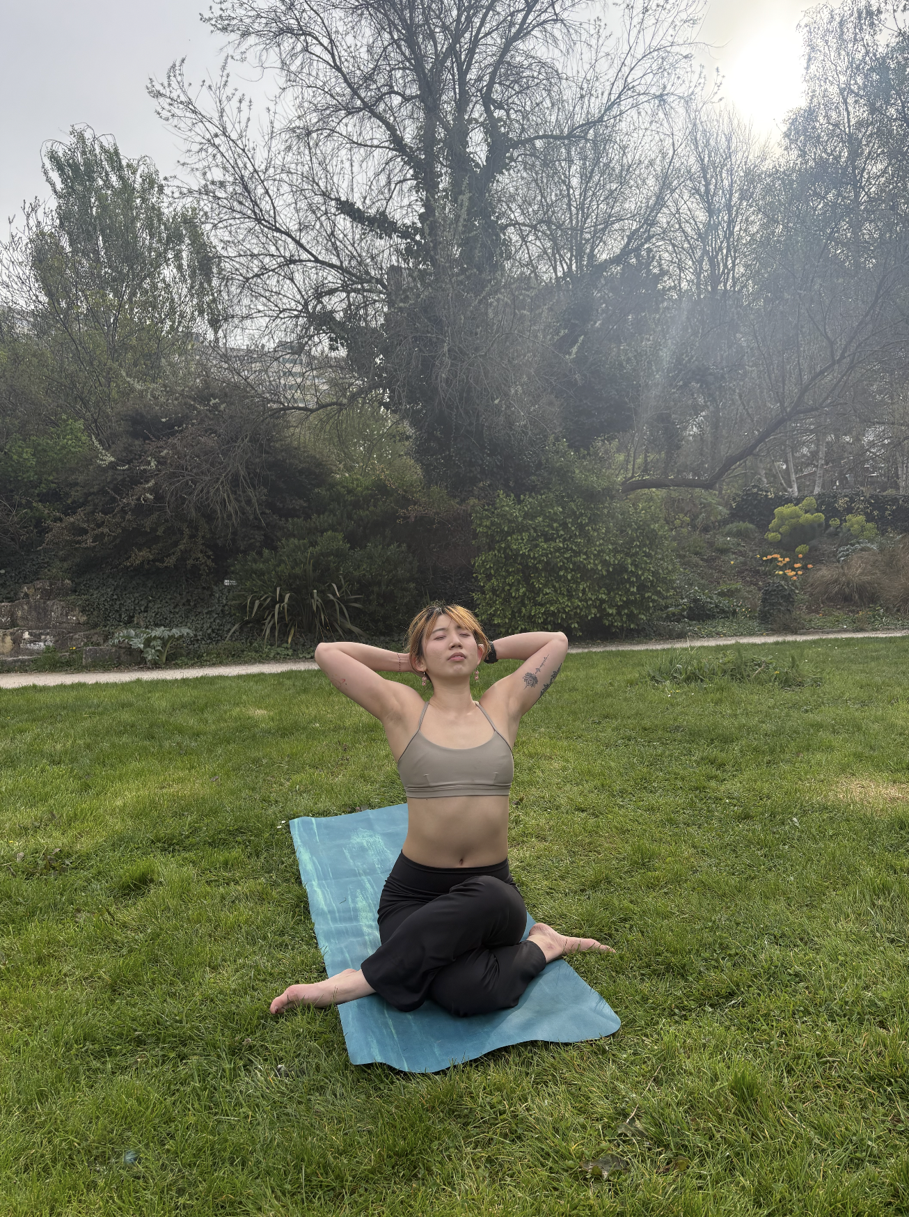 A woman practicing yoga outdoors on a grassy lawn, sitting in a kneeling pose with arms behind her head and eyes closed, with trees and bushes in the background.