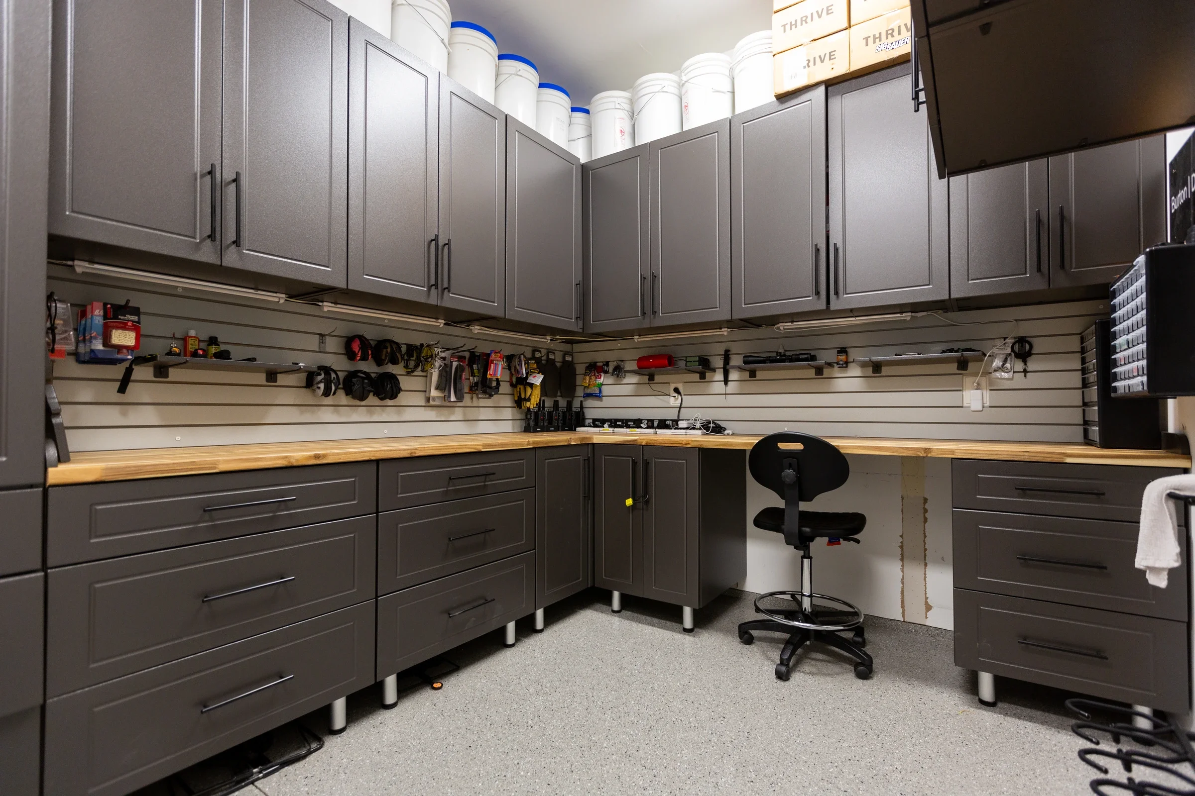 Inside a workshop or storage room with gray cabinets, a workbench, and a black swivel chair. Items such as tools, helmets, and supplies are hanging on the slatwall, with large white buckets stored on top of the cabinets and storage boxes on the right.